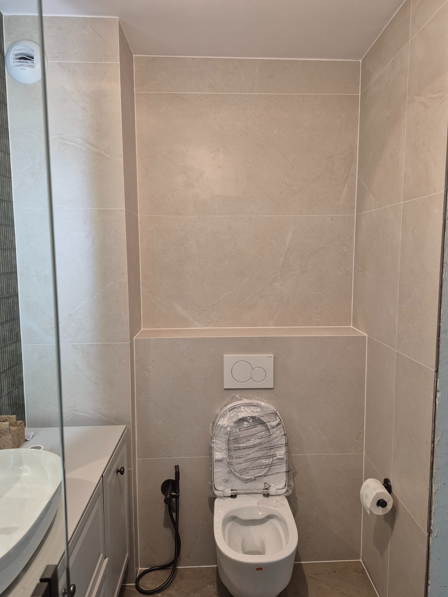 Nowoczesna, beżowa łazienka z podwieszaną toaletą w folii ochronnej, bidetką i rolką papieru. Ściany wyłożone płytkami imitującymi marmur. Widoczna umywalka i szafka.