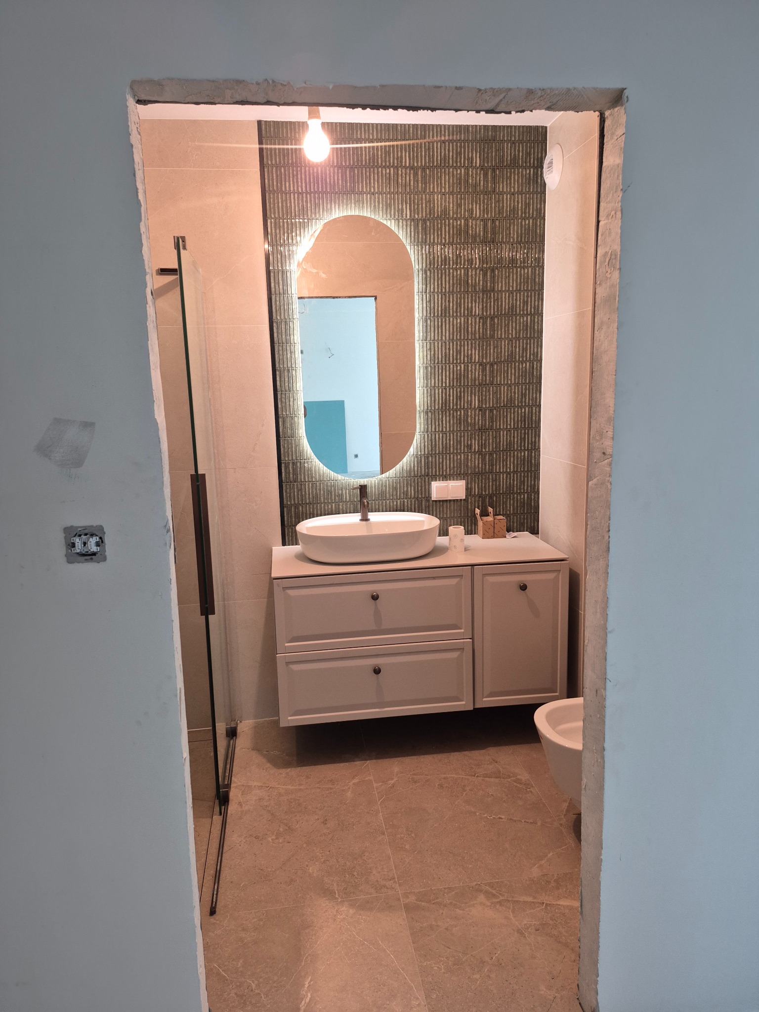 Nowoczesna łazienka z owalnym lustrem podświetlanym LED, białą umywalką nablatową i płytkami w odcieniach beżu i szarości, widziana z korytarza.