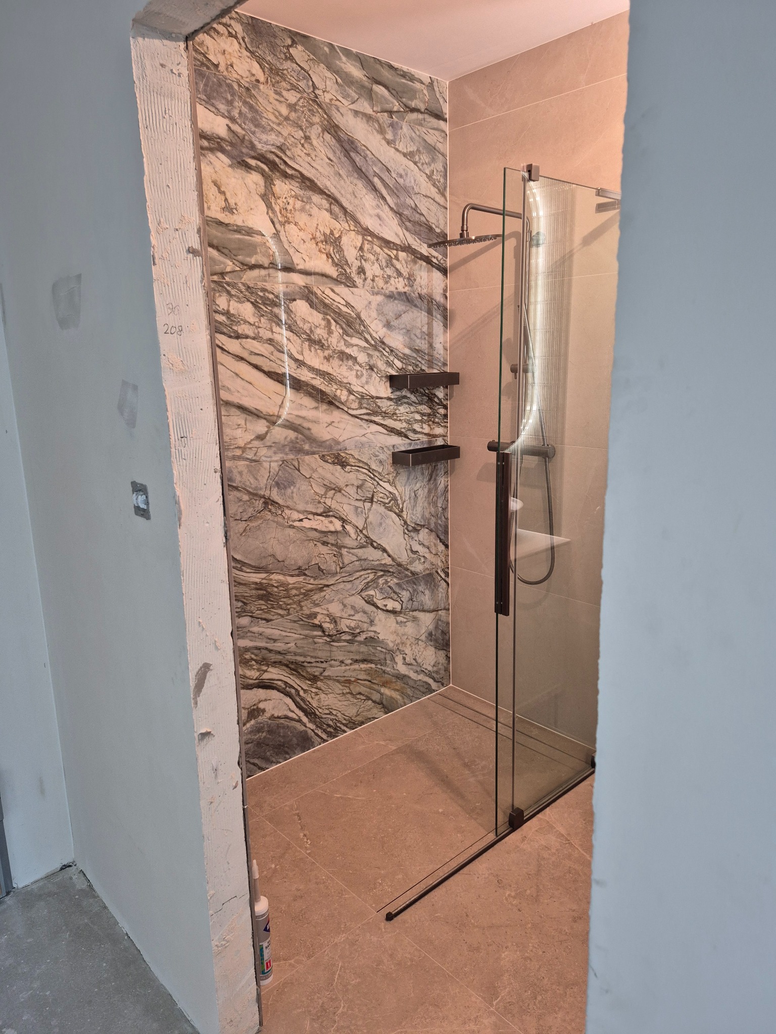 Nowoczesna kabina prysznicowa z marmurową ścianą i szklanymi drzwiami, widoczna przez otwarte drzwi w trakcie wykończenia łazienki.