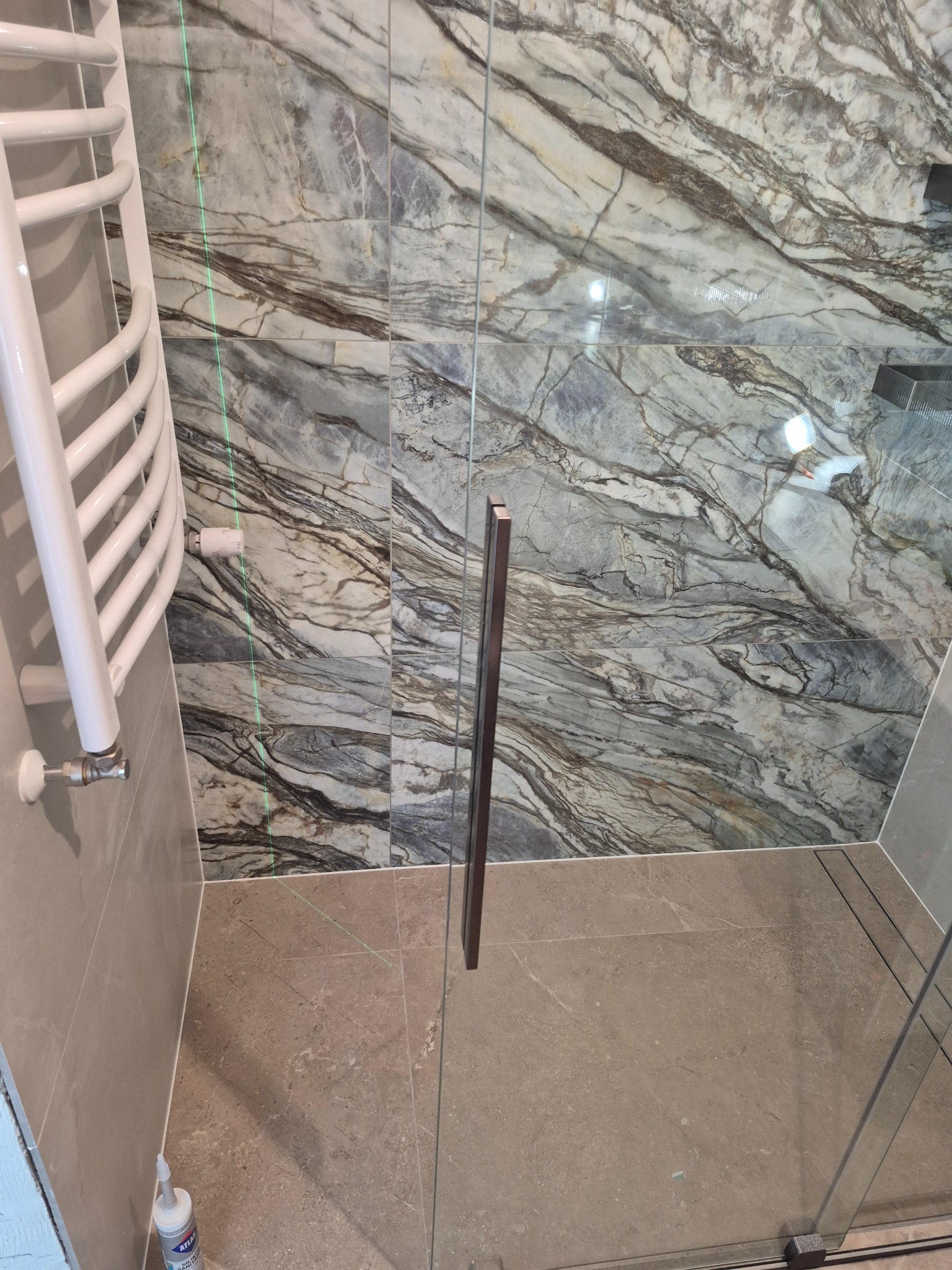 Wykończona łazienka z marmurowymi płytkami, szklaną kabiną prysznicową i białym grzejnikiem drabinkowym. Widoczna zielona linia lasera.