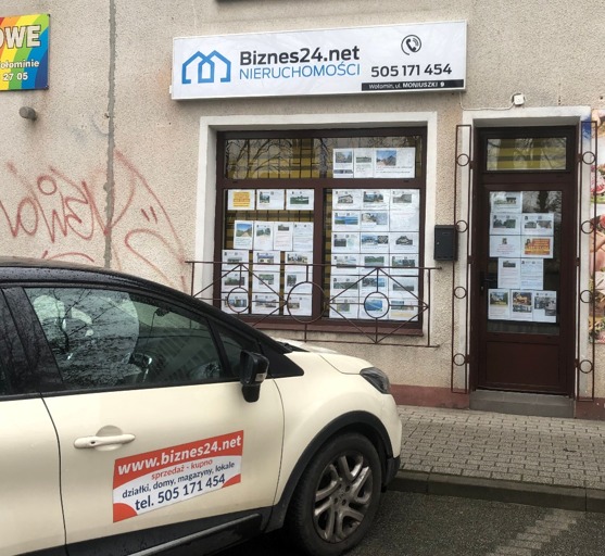 Fasada biura nieruchomości z szyldem 'Biznes24.net' i ogłoszeniami w oknach, obok zaparkowany samochód z naklejką reklamową tej samej firmy.