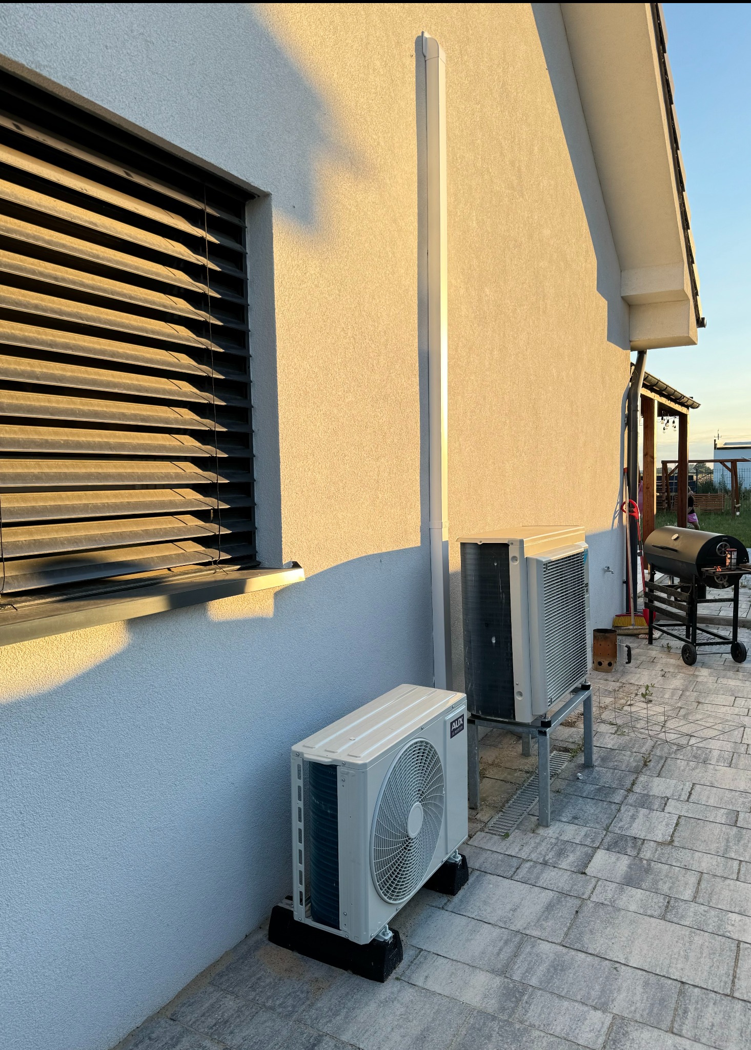Dwa klimatyzatory typu split zamontowane na zewnątrz budynku jednorodzinnego, model AUX Q-Smart Plus widoczny na jednym z urządzeń, na kostce brukowej, w tle grill.