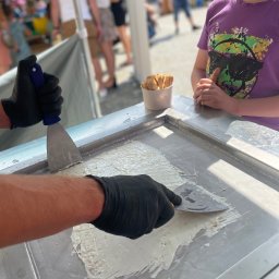 Gastrolandia Bartosz Urbanek - Przygotowywanie lodów tajskich na metalowej płycie chłodzącej. Widoczne dłonie w czarnych rękawiczkach formujące masę szpachelkami. Obok stoi dziecko obserwujące proces.