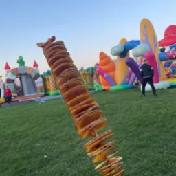 Spiralne chipsy ziemniaczane na patyku, trzymane w dłoni w czarnej rękawiczce, w tle dmuchane zamki i zielony trawnik.