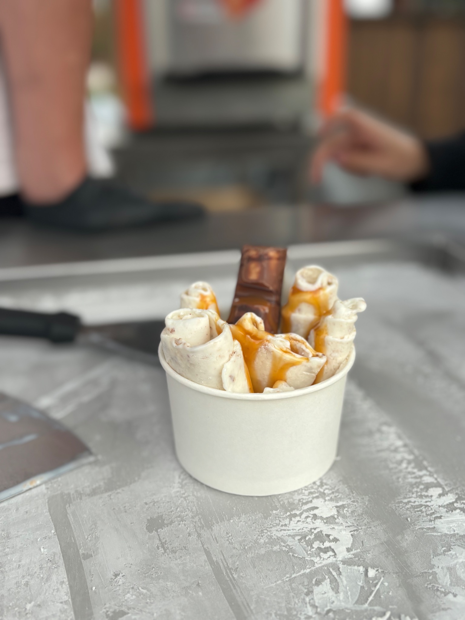 Porcja lodów tajskich w kubku, polana karmelem i z kawałkiem czekolady, na metalowej płycie do przygotowywania lodów.