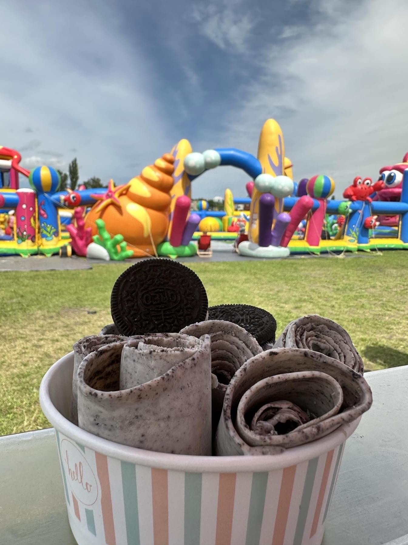 Rolowane lody Oreo w papierowym kubku z napisem 'hello', na tle dmuchanego placu zabaw w plenerze.