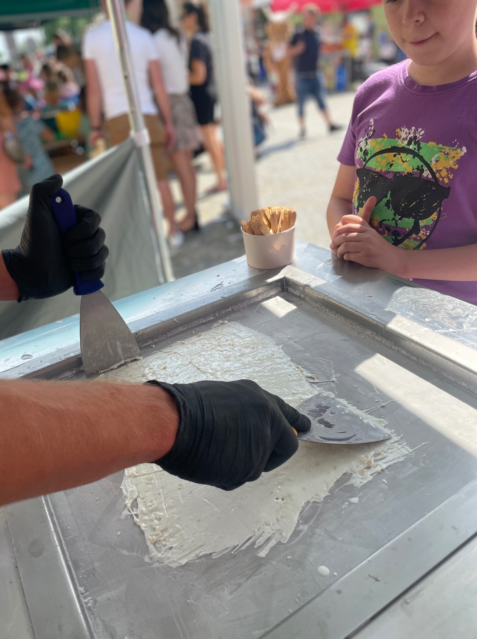 Przygotowywanie lodów tajskich na metalowej płycie chłodzącej. Widoczne dłonie w czarnych rękawiczkach formujące masę szpachelkami. Obok stoi dziecko obserwujące proces.