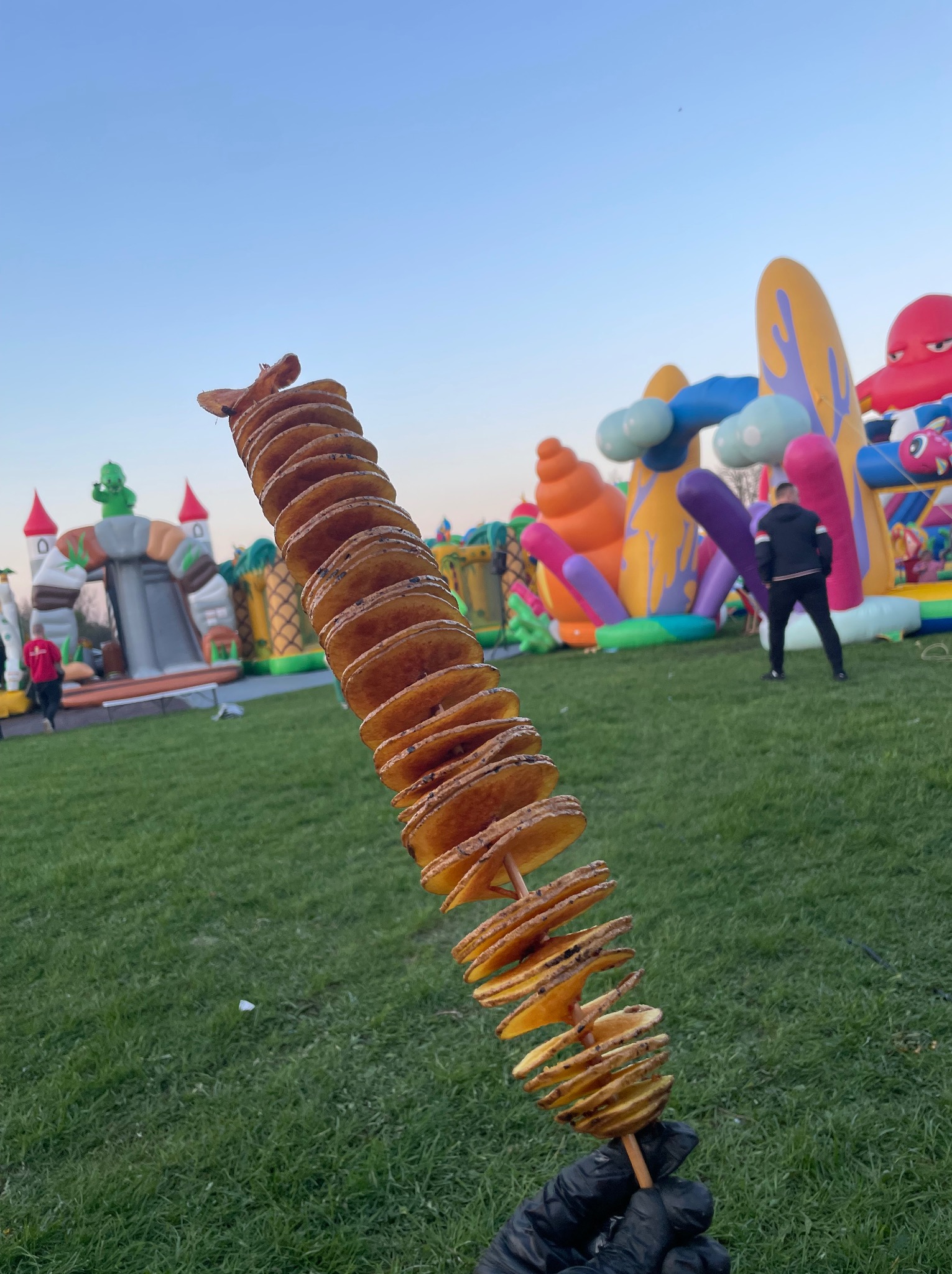 Spiralne chipsy ziemniaczane na patyku, trzymane w dłoni w czarnej rękawiczce, w tle dmuchane zamki i zielony trawnik.