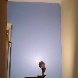 PBSKR.BUD - Niebieska ściana po malowaniu, widoczna z dołu z lampą roboczą na drabinie. Fragment futryny po lewej, żółta ściana po prawej. Świeżo pomalowana.