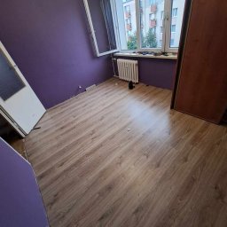 PBSKR.BUD - Pokój po remoncie: świeżo pomalowane fioletowe ściany, nowe panele podłogowe imitujące drewno, biały grzejnik pod oknem z widokiem na budynek.