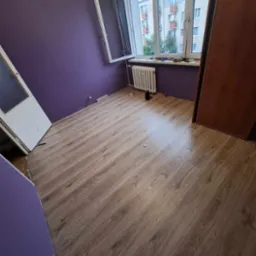 Pokój po remoncie: świeżo pomalowane fioletowe ściany, nowe panele podłogowe imitujące drewno, biały grzejnik pod oknem z widokiem na budynek.