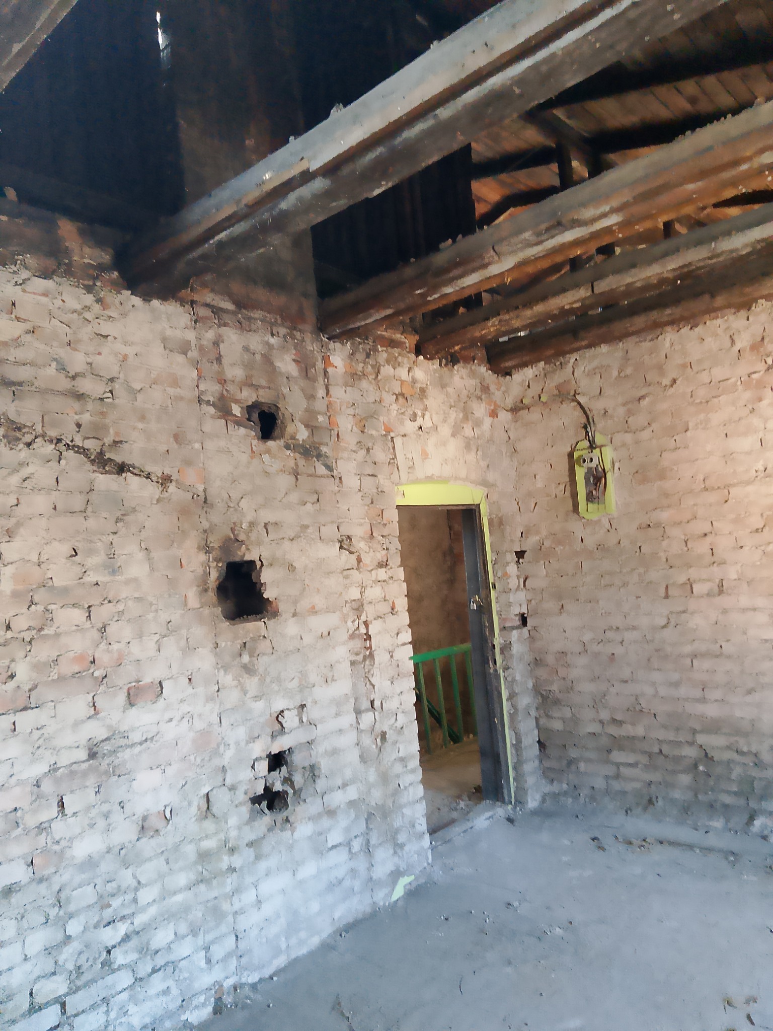 Wnętrze budynku w trakcie prac remontowych: odsłonięta ceglana ściana z otworami, drewniane belki stropowe, stara instalacja elektryczna i zielona balustrada w przejściu.