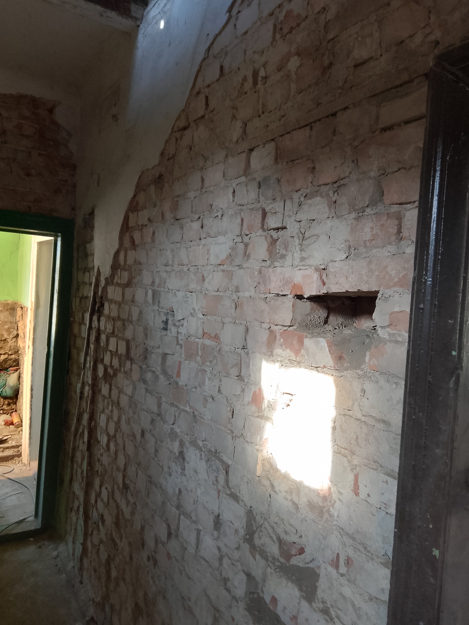 Fragment ściany z czerwonej cegły z otworem, z widocznym światłem słonecznym wpadającym do wnętrza, obok ciemna framuga drzwi.