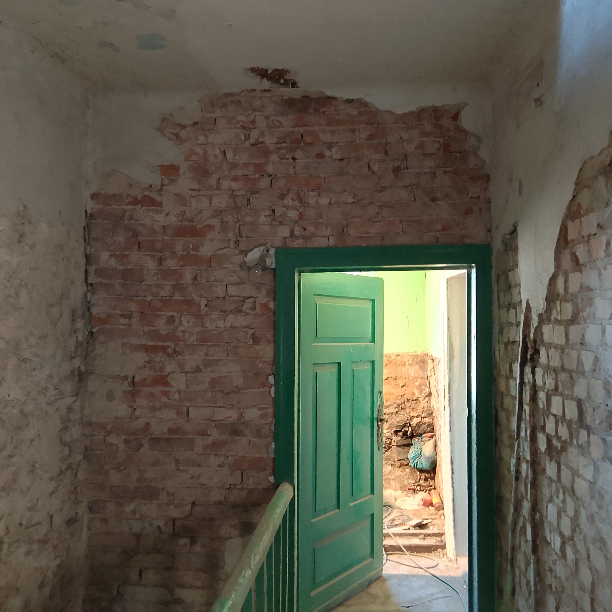 Wnętrze w trakcie renowacji: odsłonięta cegła na ścianie, zielone drzwi z widocznymi śladami użytkowania, fragment schodów i gruz w sąsiednim pomieszczeniu.