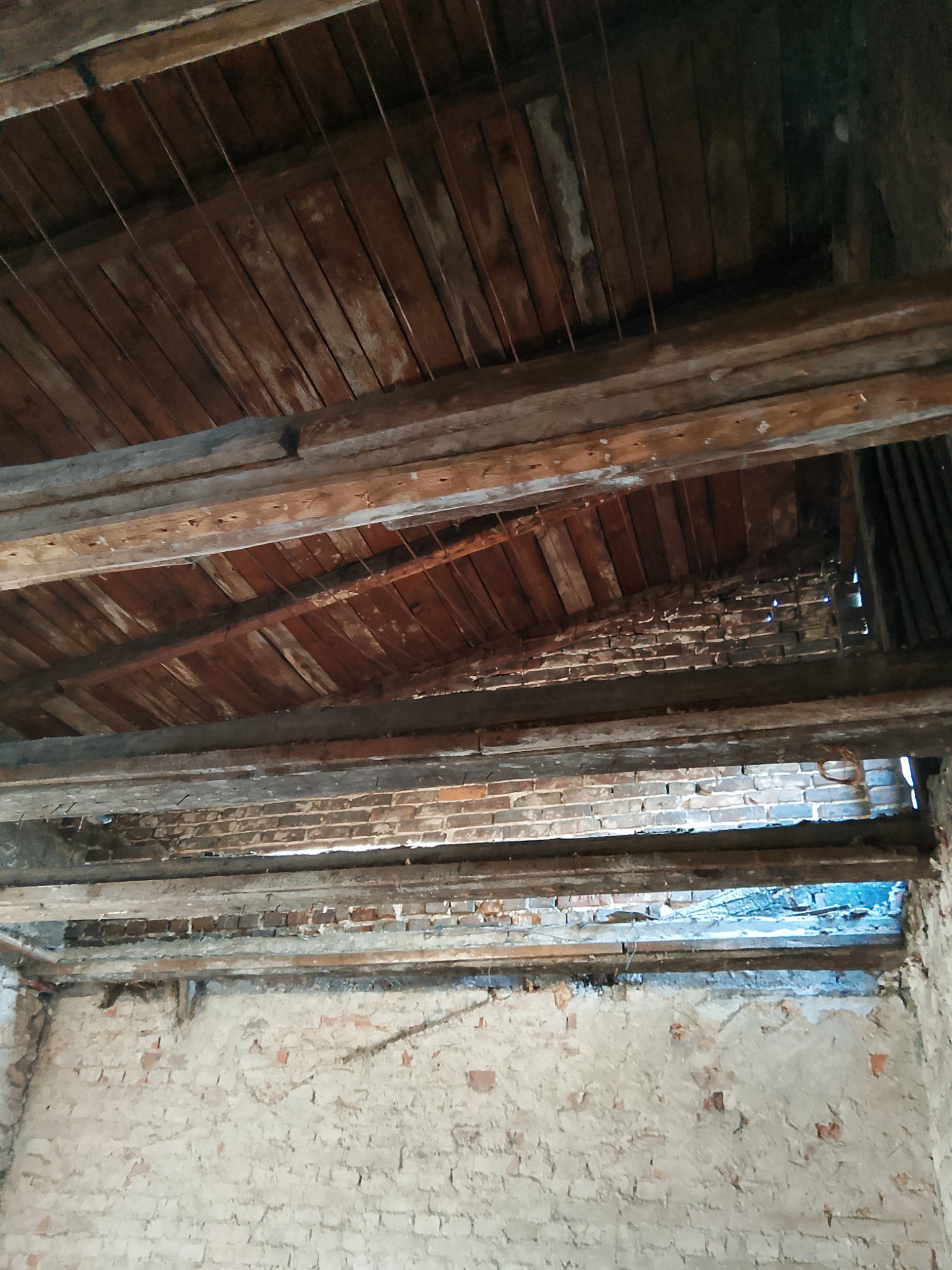 Wnętrze starego budynku z odsłoniętym drewnianym stropem i fragmentami muru ceglanego, widoczne ślady po usuniętych ścianach działowych.