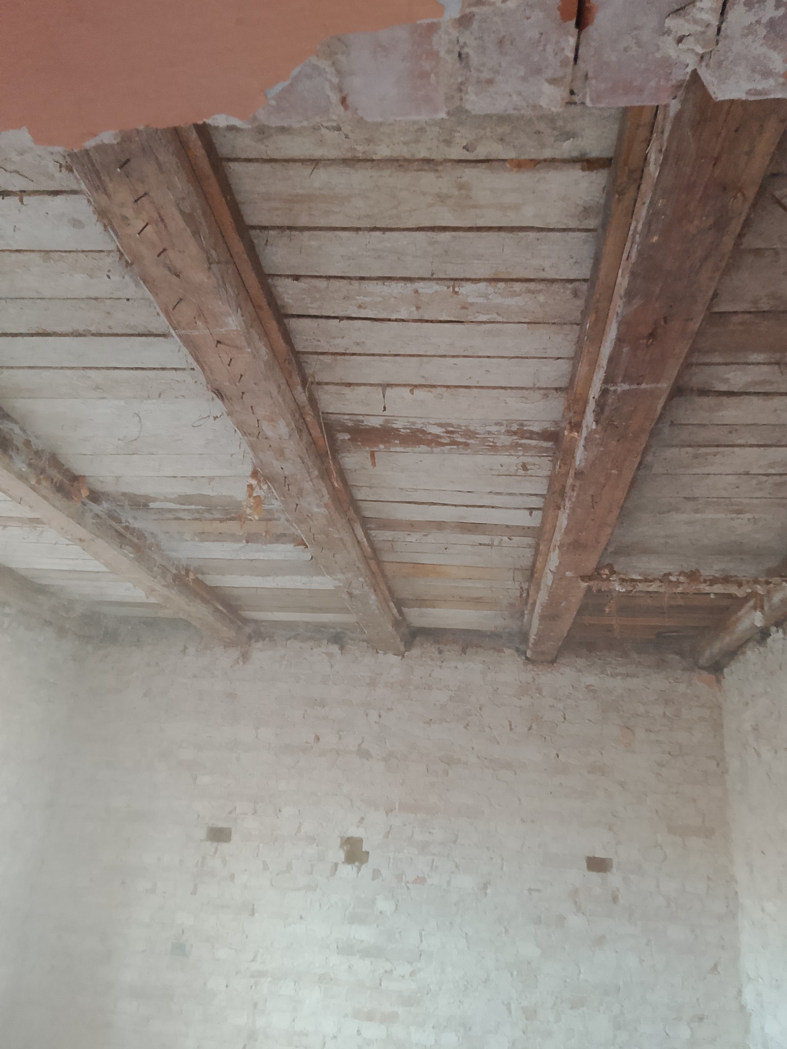 Fragment zdemontowanego drewnianego stropu z widocznymi belkami nośnymi i deskami poszycia, odsłonięta ceglana ściana z ubytkami tynku.
