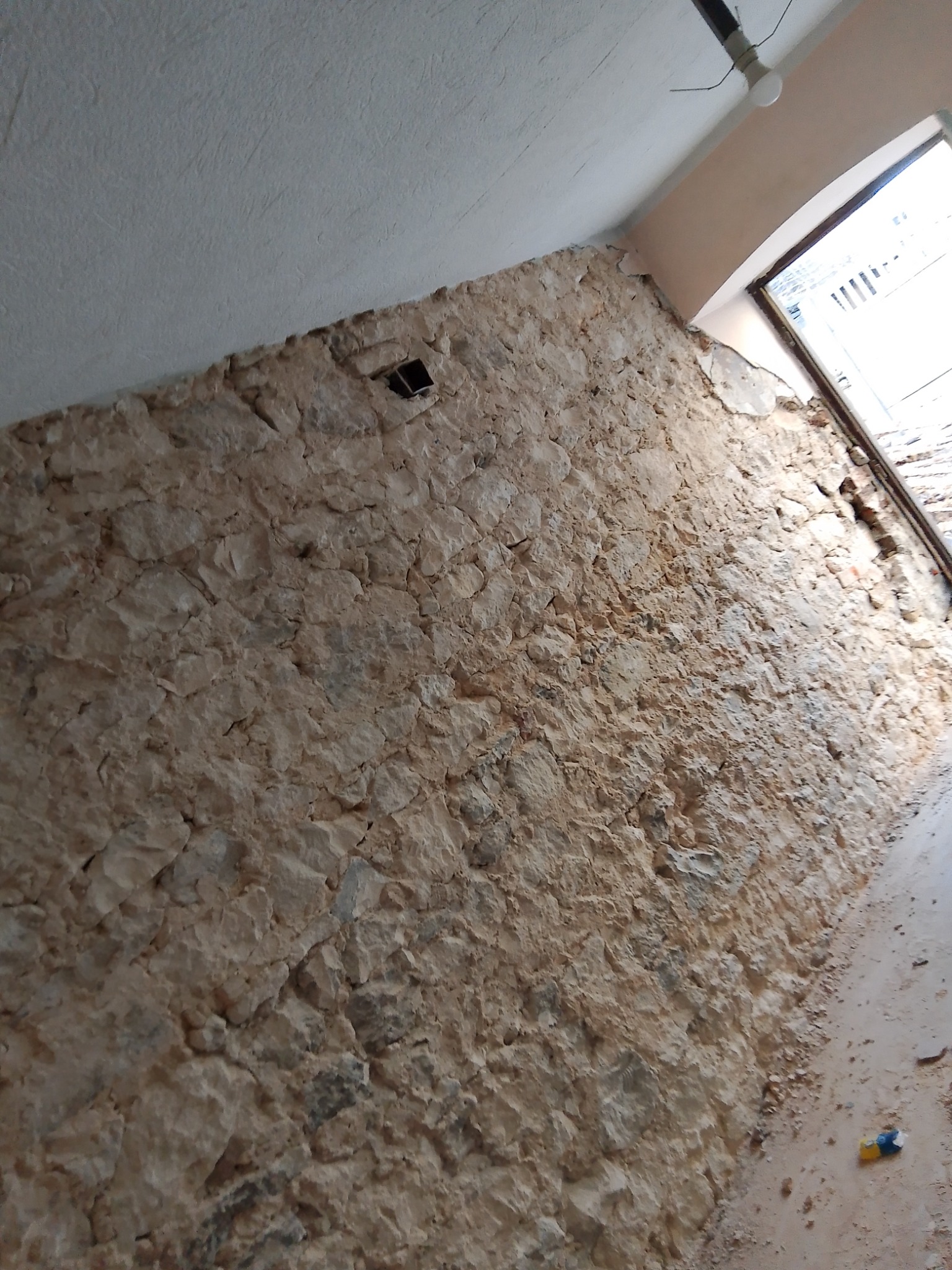 Fragment ściany z kamienia wapiennego z widocznym otworem wentylacyjnym, surowy stan przed wykończeniem, widoczne ślady prac budowlanych i okno w tle.