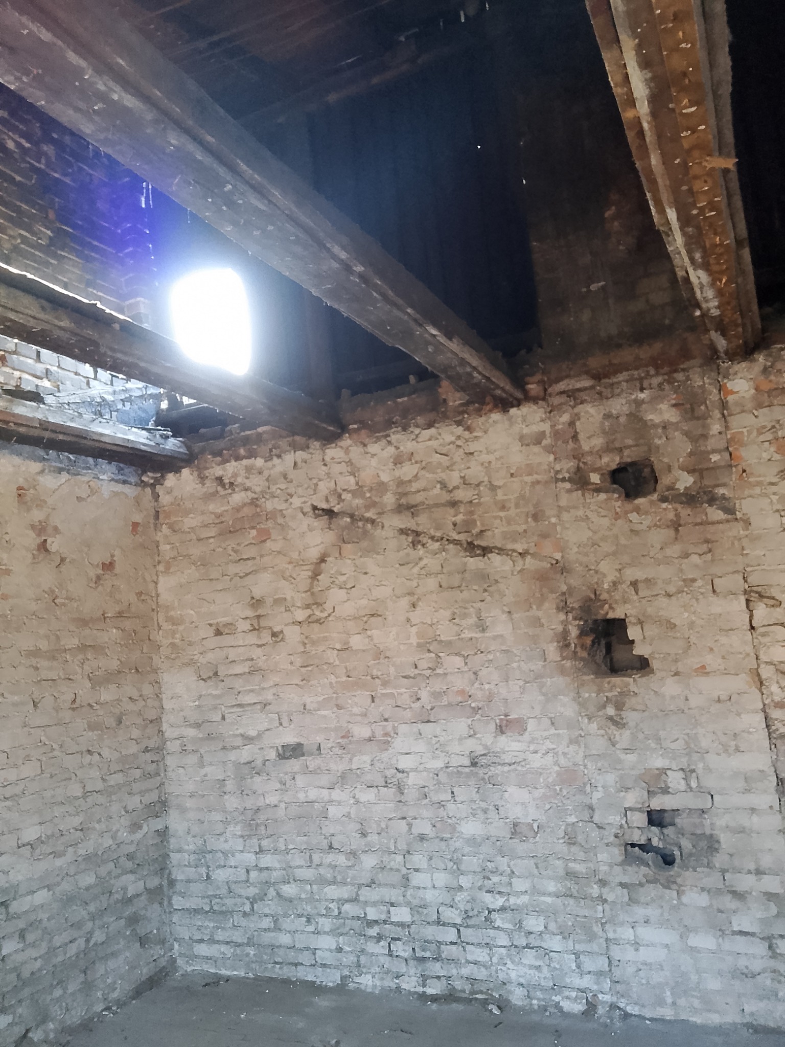 Wnętrze starego, ceglanego budynku w trakcie rozbiórki, z widocznymi fragmentami drewnianej konstrukcji stropu i otworami w ścianach, oświetlone naturalnym światłem wpadającym przez otwór w dachu.