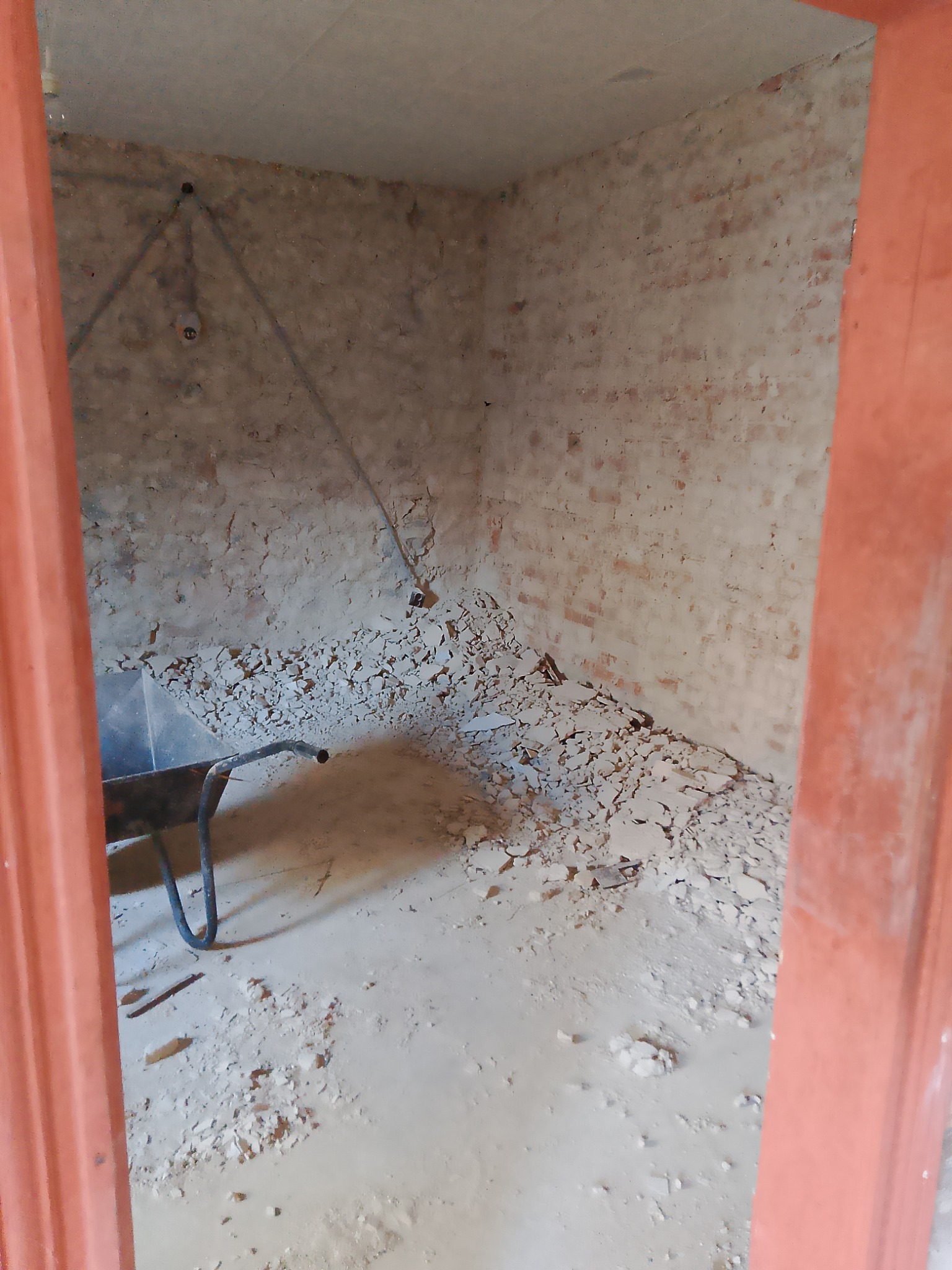 Wnętrze pomieszczenia w trakcie remontu, z gruzem i fragmentami ściany na podłodze, widoczna taczka i fragment instalacji na ścianie.