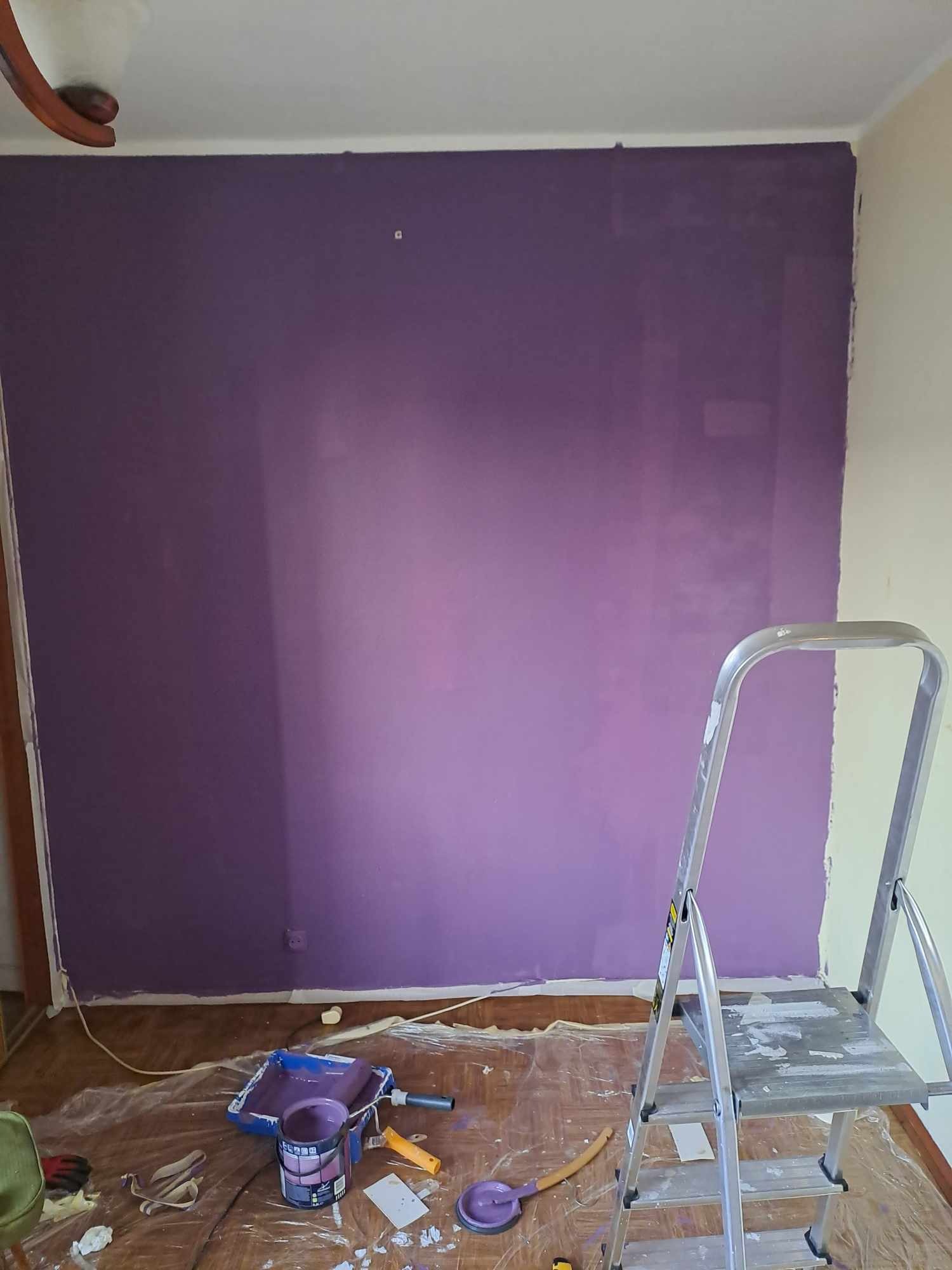Ściana pomalowana na fioletowo w trakcie malowania. Widoczne narzędzia malarskie, drabina, podłoga zabezpieczona folią. Świeża, nierównomierna warstwa farby.