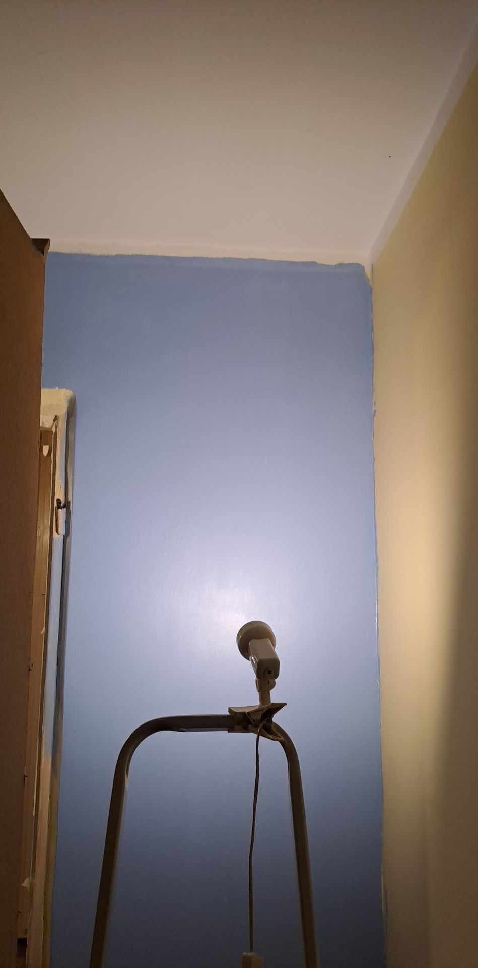 Niebieska ściana po malowaniu, widoczna z dołu z lampą roboczą na drabinie. Fragment futryny po lewej, żółta ściana po prawej. Świeżo pomalowana.