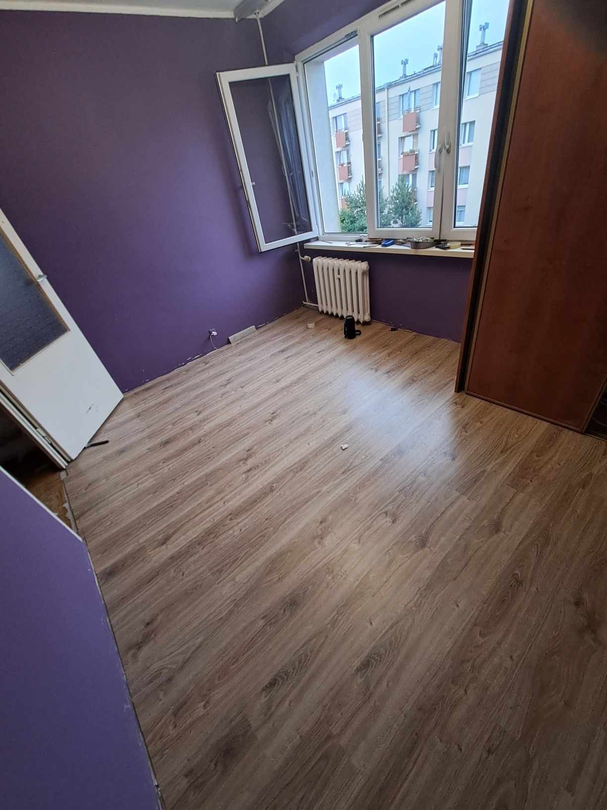 Pokój po remoncie: świeżo pomalowane fioletowe ściany, nowe panele podłogowe imitujące drewno, biały grzejnik pod oknem z widokiem na budynek.
