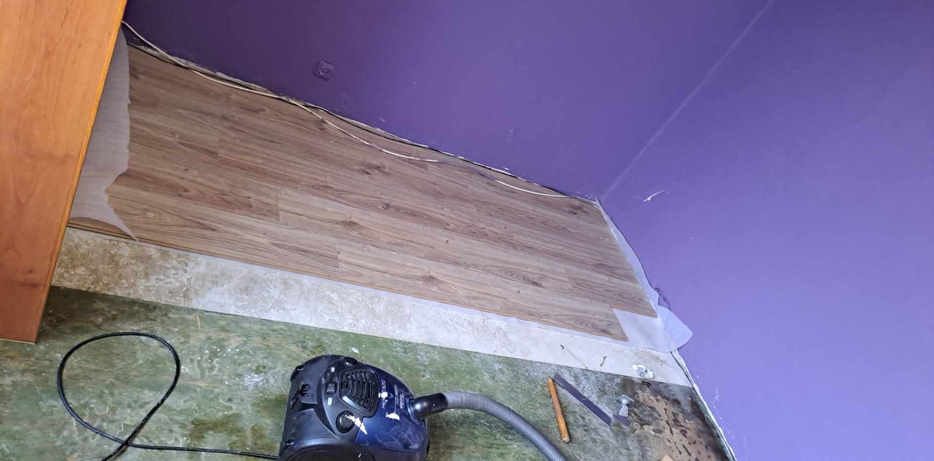 Układanie paneli podłogowych w pokoju z fioletowymi ścianami. Widoczny odkurzacz i częściowo położona podłoga.