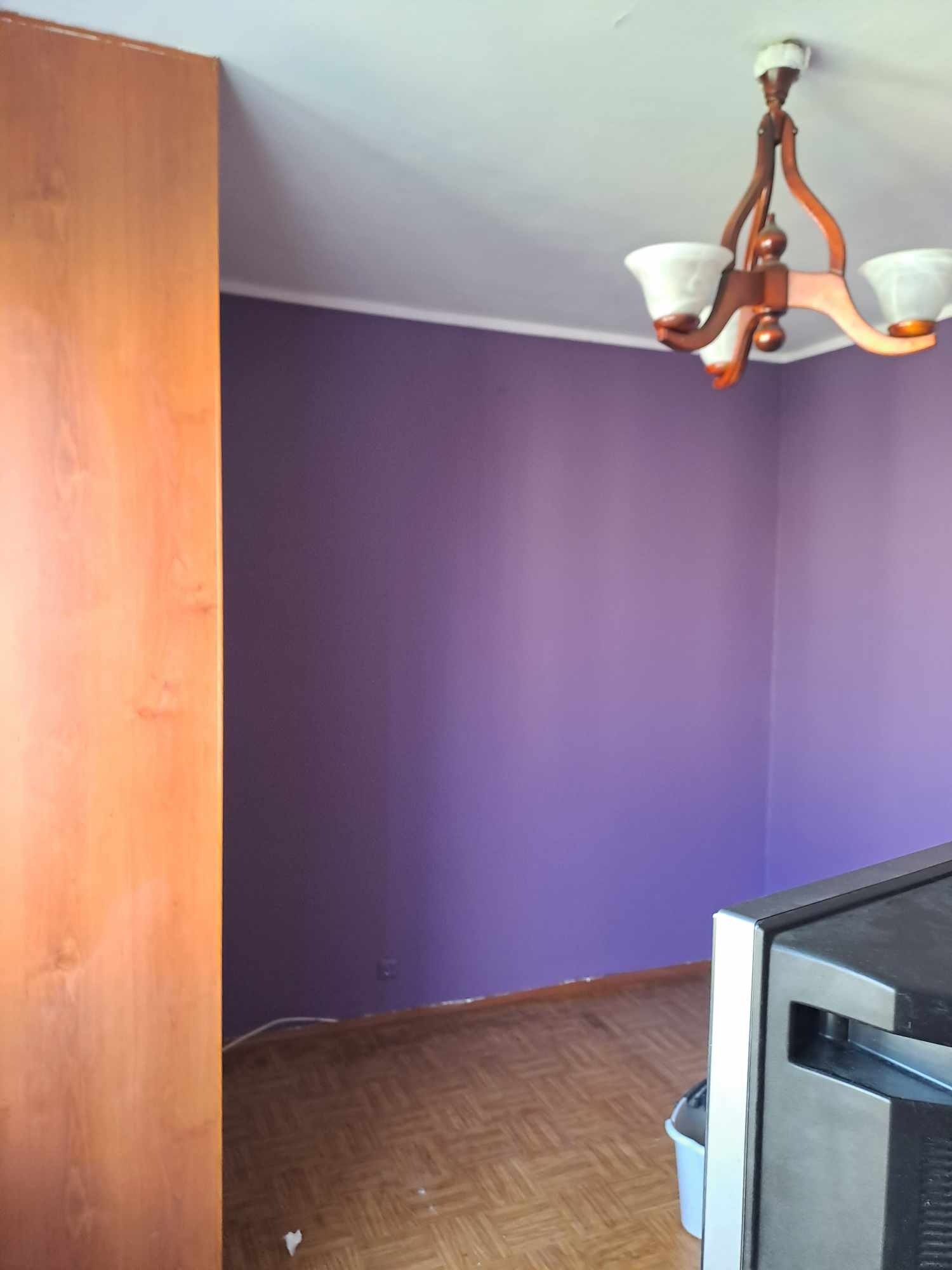Fioletowa ściana w pokoju z drewnianą podłogą i lampą sufitową. Widoczny fragment szafy i sprzętu AGD. Świeżo pomalowane ściany.