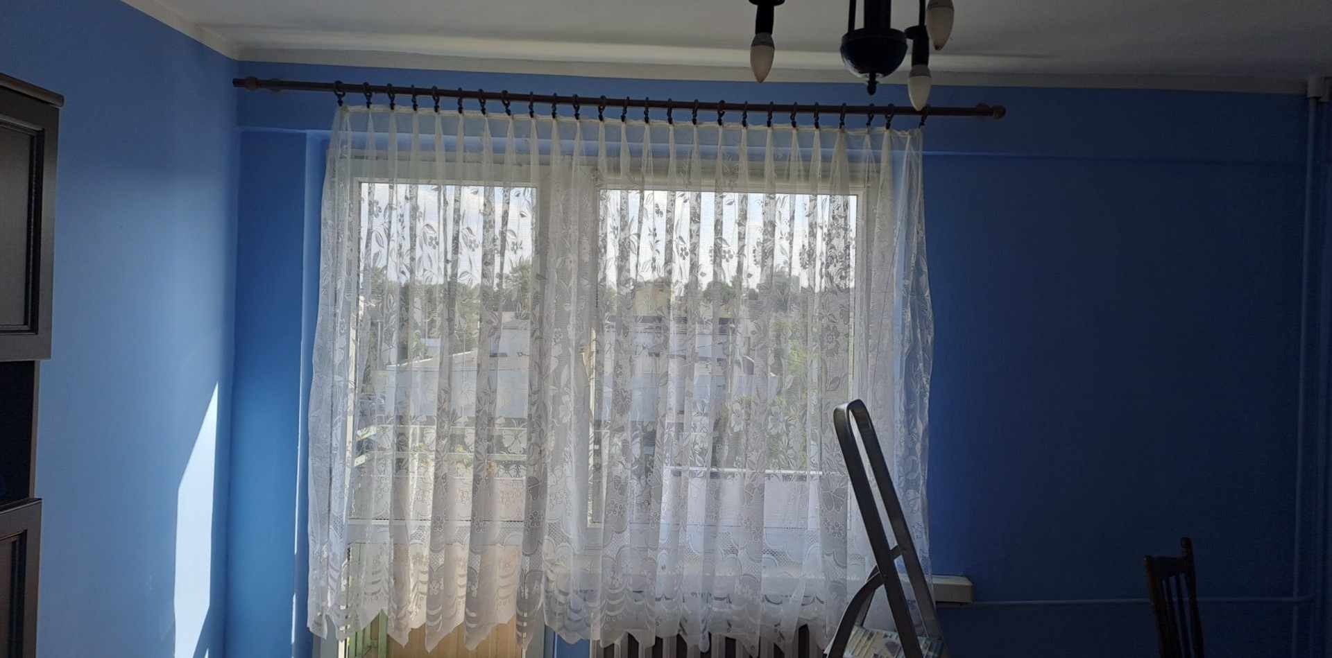 Błękitne ściany po malowaniu, okno z firanką i drabina malarska. Widoczne niedoskonałości malarskie przy oknie, podkreślone padającym światłem.