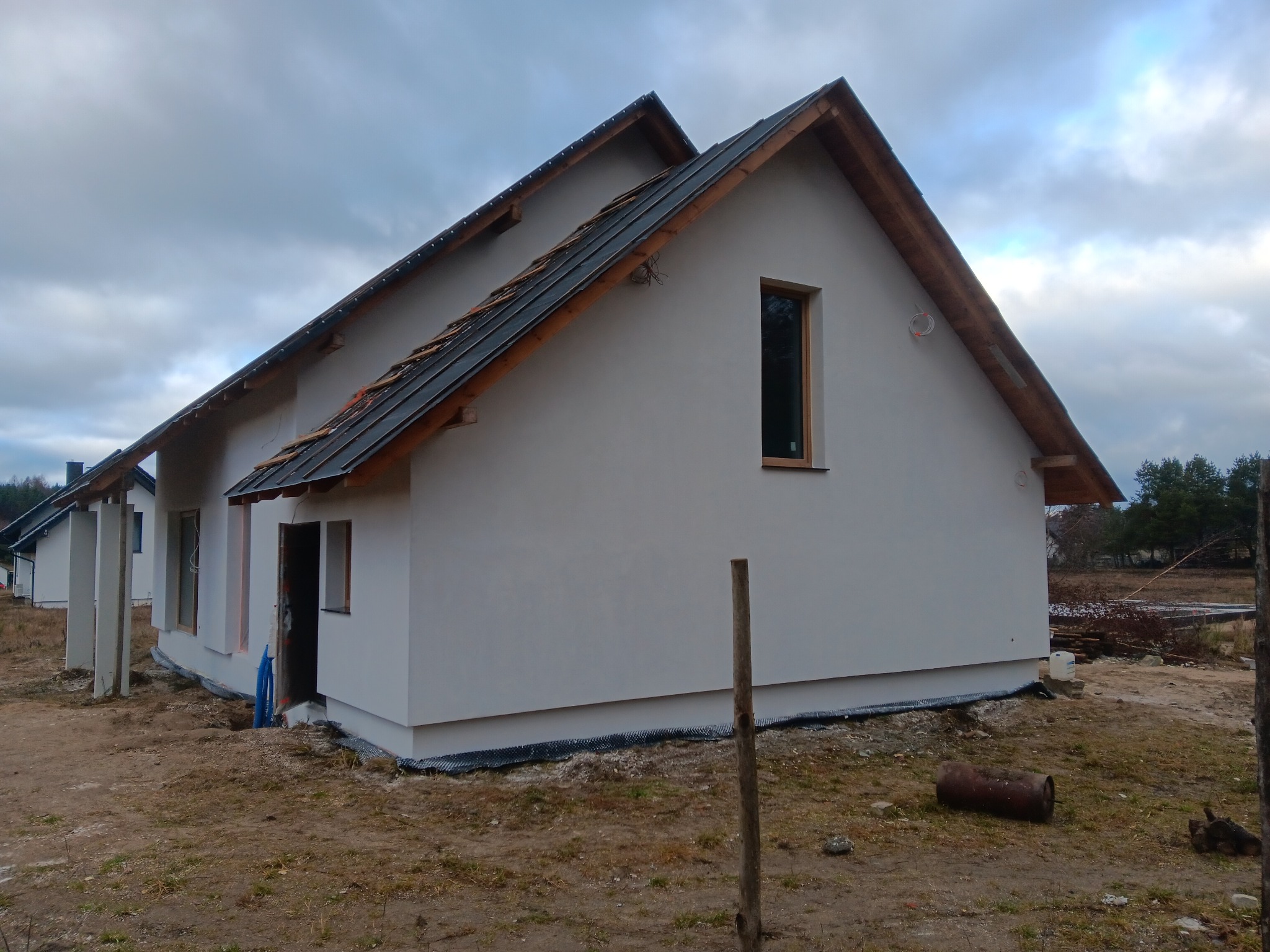 Biały dom jednorodzinny z ciemnym dachem w trakcie budowy, widoczne surowe wykończenie elewacji i otoczenia. Okno z drewnianą ramą. Ujęcie z boku, pochmurne niebo.