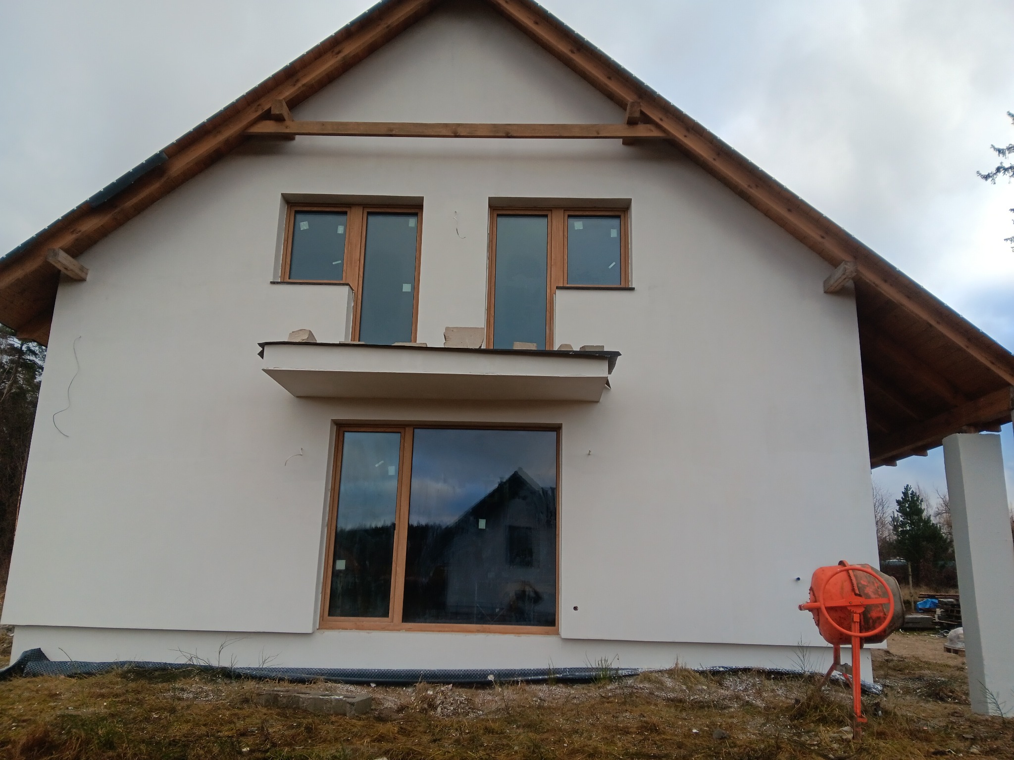 Świeżo otynkowany dom jednorodzinny z drewnianą konstrukcją dachu i oknami. Na pierwszym planie widoczna betoniarka, sugerująca trwające prace wykończeniowe.