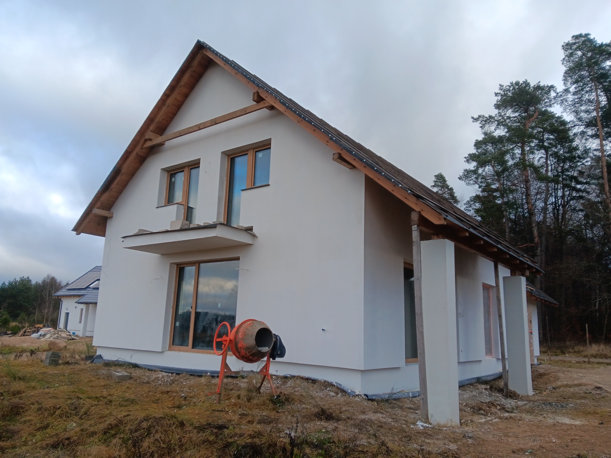 Nowy dom jednorodzinny z białą elewacją, widoczna betoniarka i fragment sąsiedniego budynku. Surowy krajobraz, pochmurne niebo.