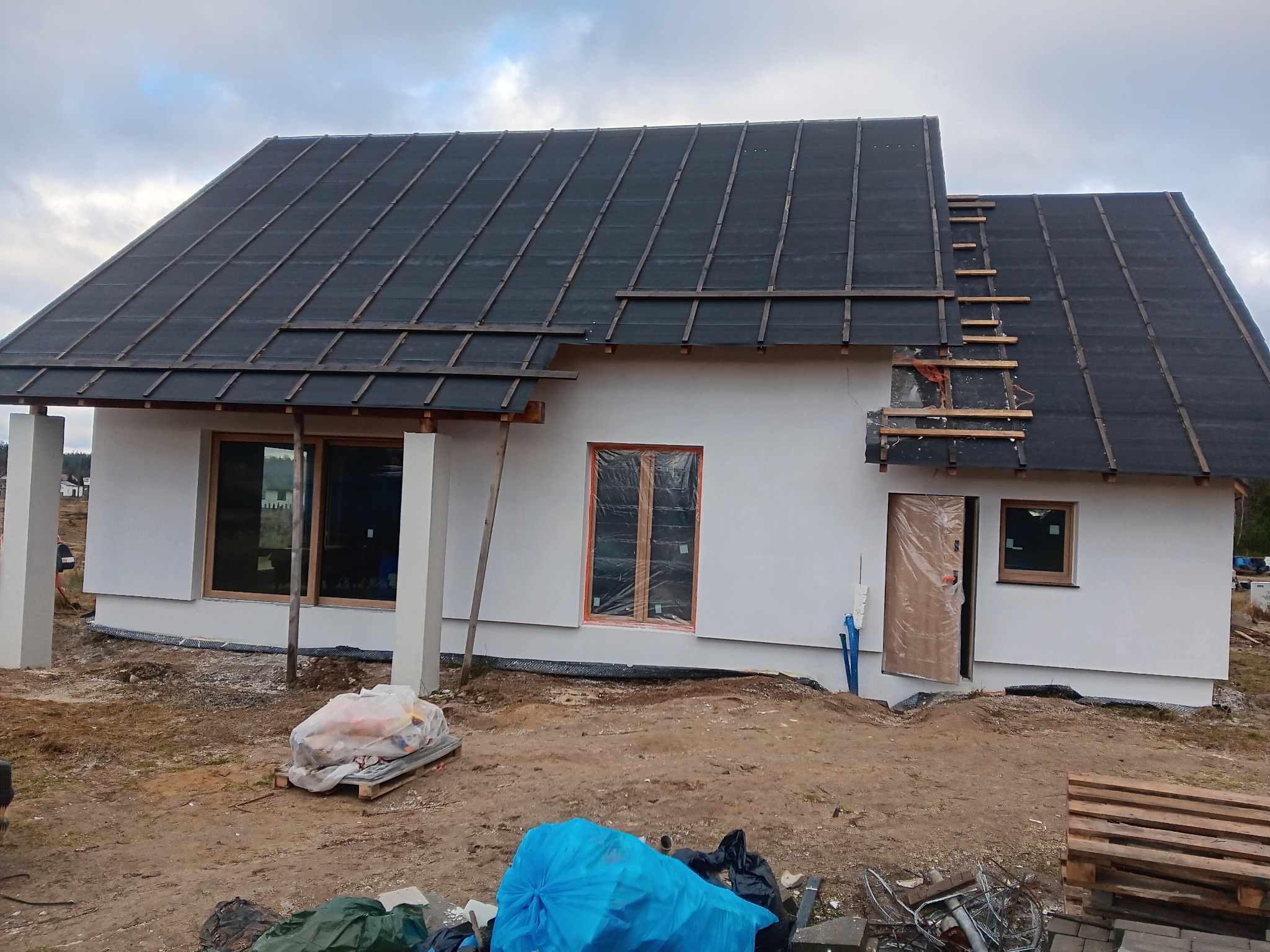 Dom jednorodzinny w trakcie budowy, z widoczną konstrukcją dachu pokrytą papą i częściowo wykonaną elewacją. Okna zabezpieczone folią, teren wokół w stanie surowym.