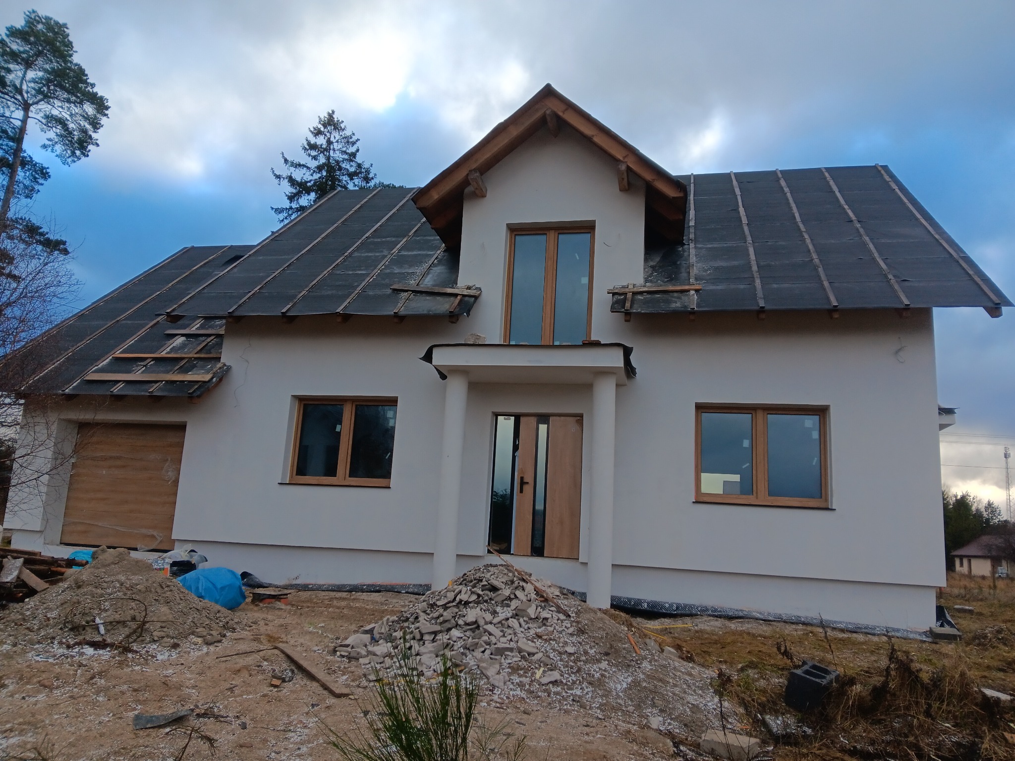 Dom jednorodzinny w trakcie budowy: biała elewacja, częściowo pokryty dach, gruz przed wejściem. Widoczne okna i drzwi. Prace budowlane w toku.