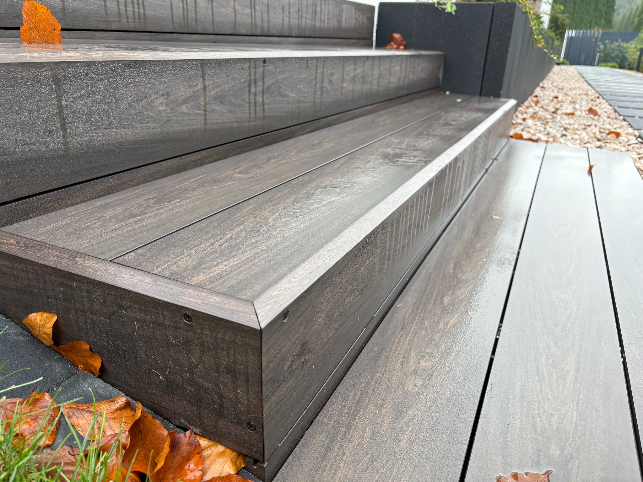 Schody z desek w kolorze ciemnego brązu, prowadzące z tarasu do ogrodu, z widocznymi śrubami mocującymi i opadłymi liśćmi.