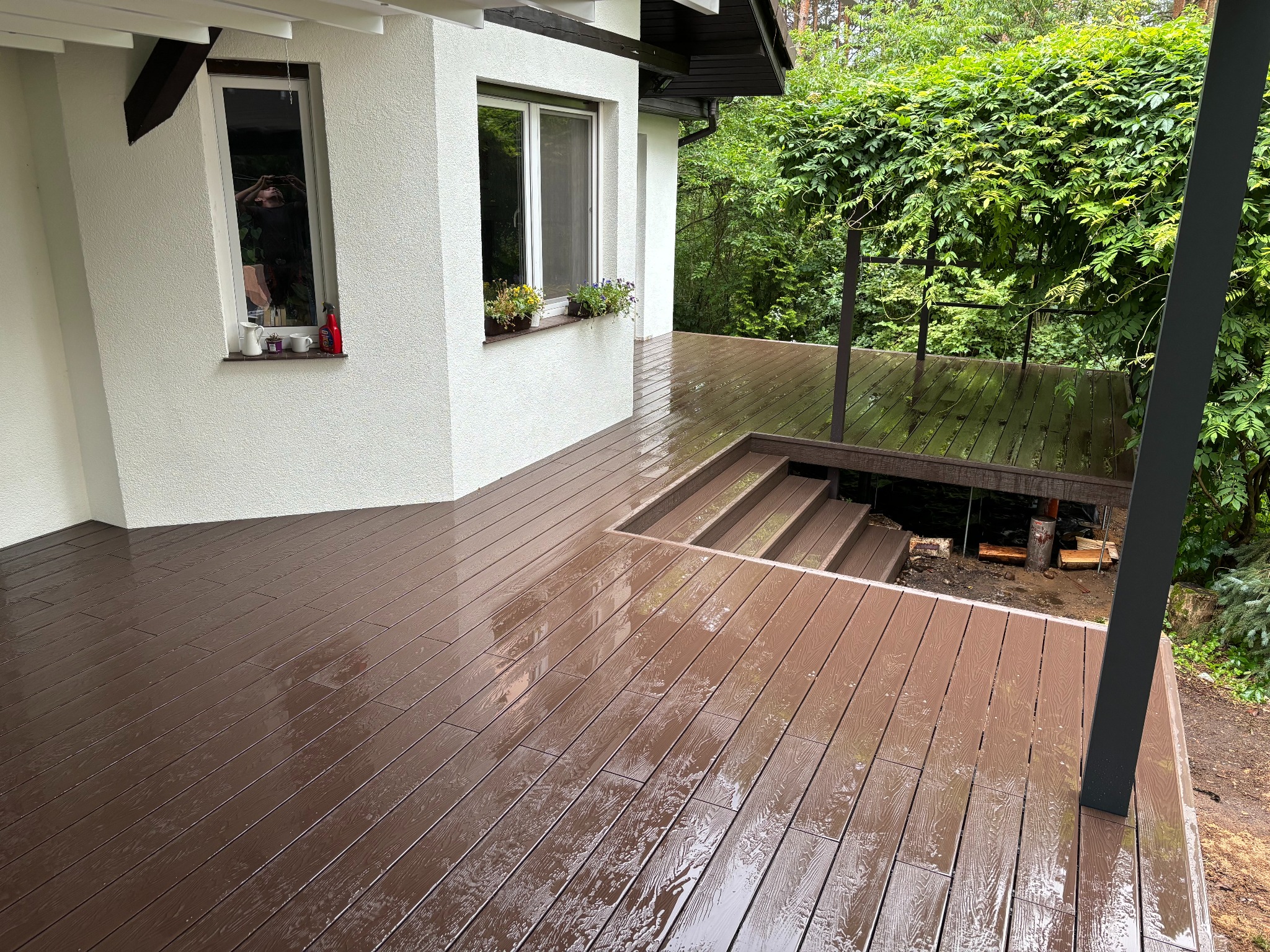 Nowoczesny taras z brązowych desek z widocznymi schodami i zadaszeniem porośniętym zielenią; widoczne odbicia na mokrej powierzchni.