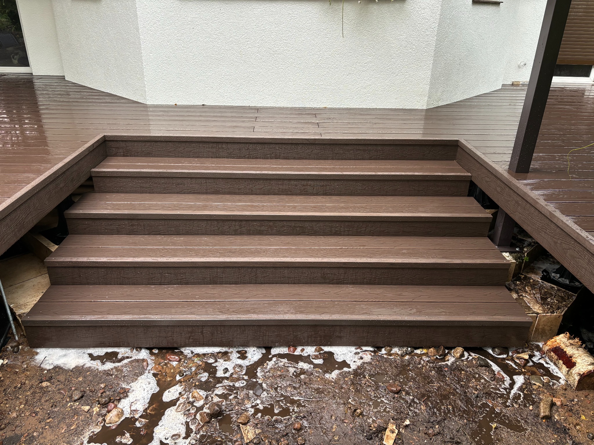 Schody z brązowych desek prowadzące na taras, widoczne ślady niedawnych prac ziemnych i błoto.