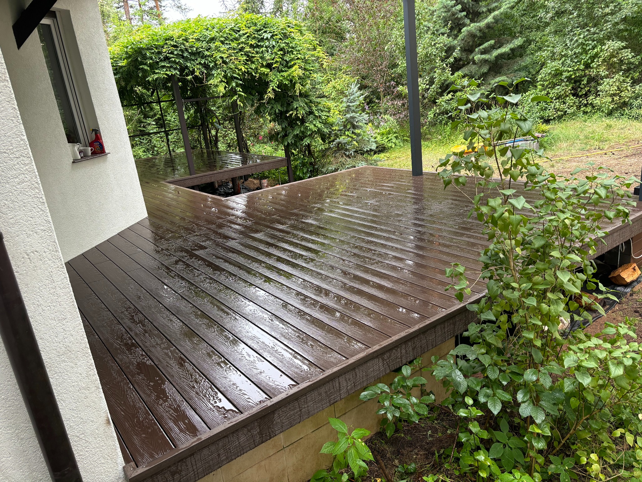 Świeżo wyczyszczony, ciemnobrązowy taras z desek kompozytowych, z widocznymi kroplami wody, przylegający do białego budynku z oknem i otoczony zielenią.