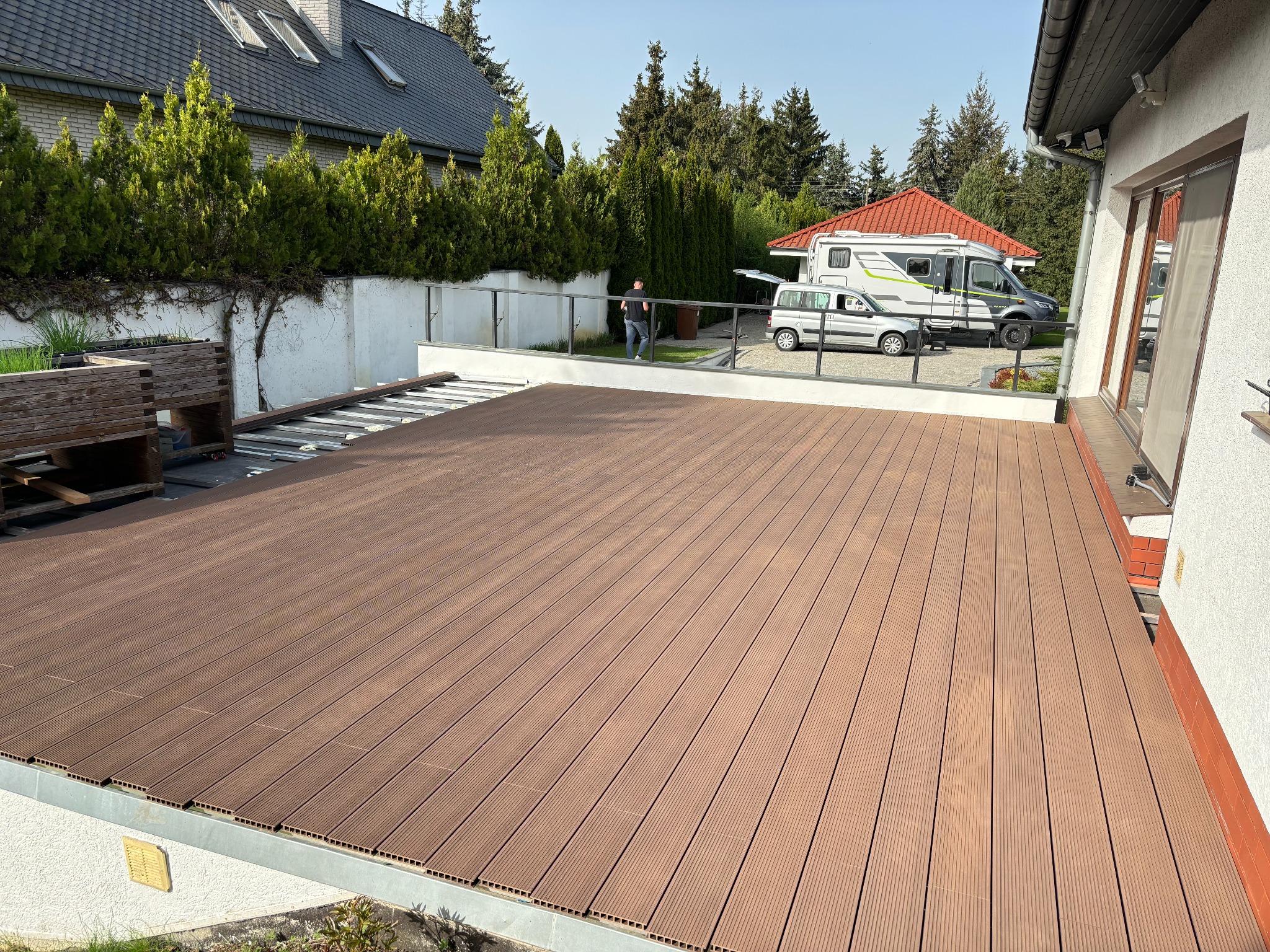 Montaż brązowego tarasu z desek na zewnątrz domu, widoczny częściowo ułożony taras, w tle zaparkowany kamper i osoba przechodząca obok.