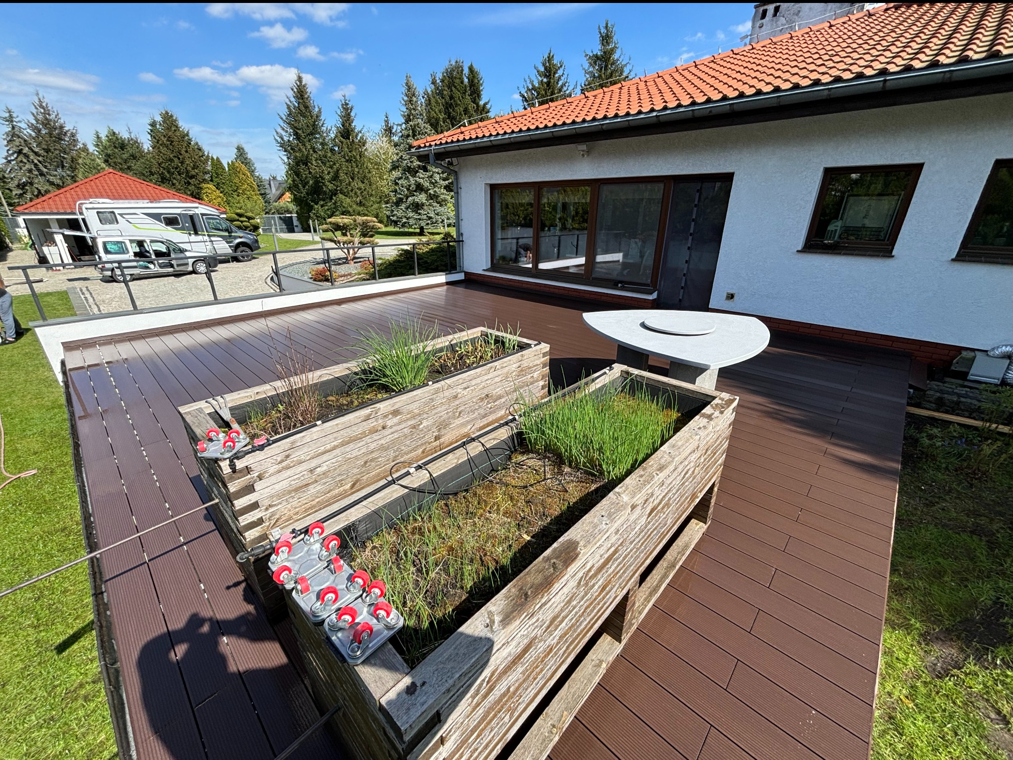 Kompozytowy taras w kolorze ciemnego brązu z drewnianymi skrzyniami na rośliny, widok na dom z dużymi oknami i teren zielony z drzewami.