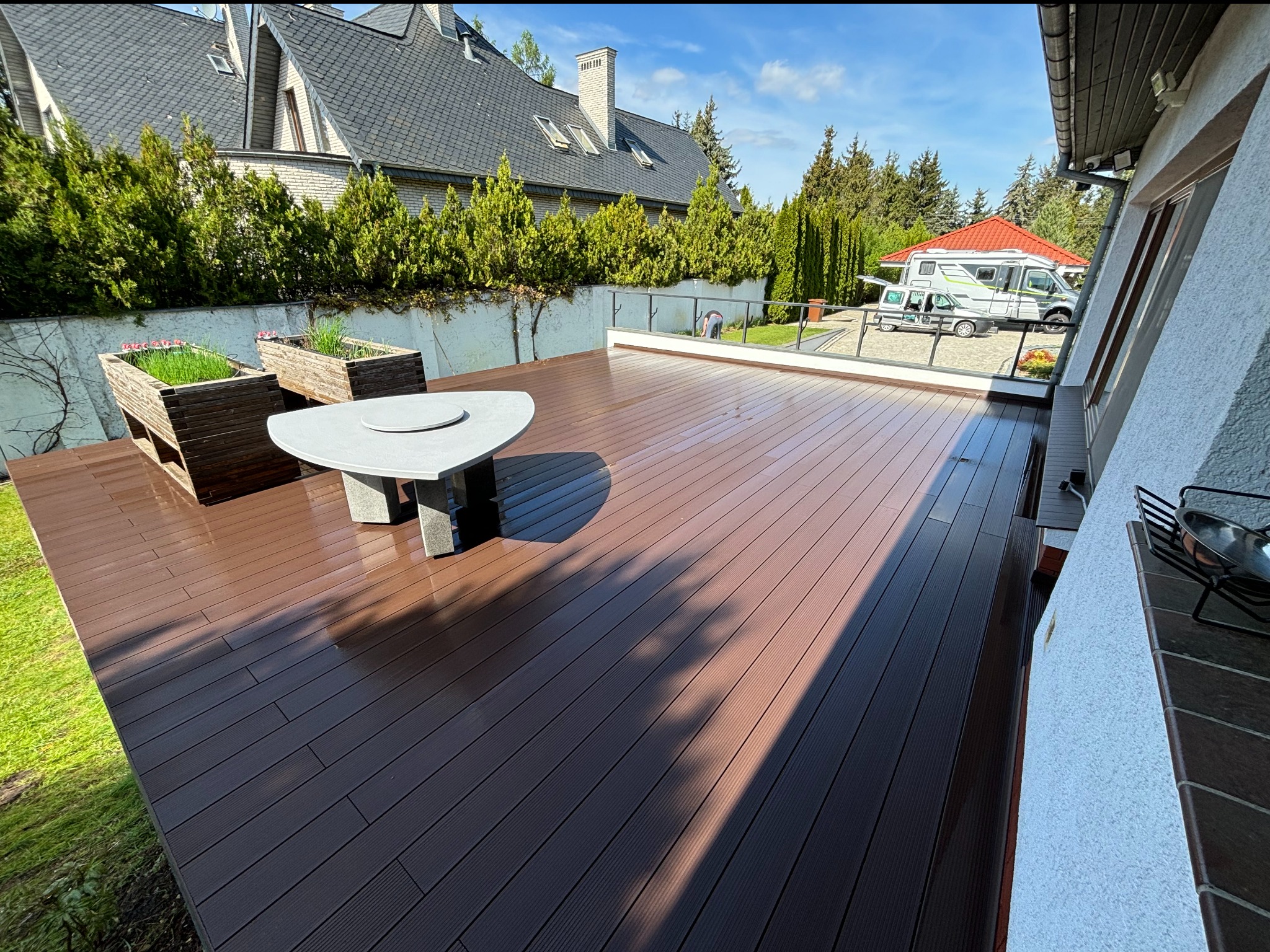 Nowoczesny taras z brązowych desek kompozytowych z minimalistycznym, trójkątnym stolikiem i donicami z zielenią, na tle domu z ogrodem i zaparkowanym kamperem.