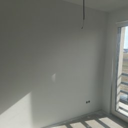 Oferteo s.a - Świeżo pomalowana ściana w jasnym odcieniu szarości z widocznymi dwoma otworami na gniazdka elektryczne i zwisającym kablem, balkon z metalową barierką i widokiem na wieś z kościołem w tle.