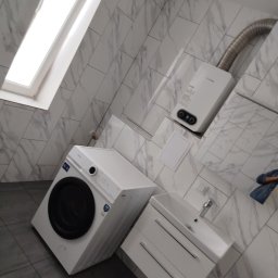 Oferteo s.a - Wykończona łazienka w bieli: pralka, szafka z umywalką, bojler i okno dachowe. Marmurowe płytki na ścianach i szare na podłodze. Nowoczesne i funkcjonalne wnętrze.
