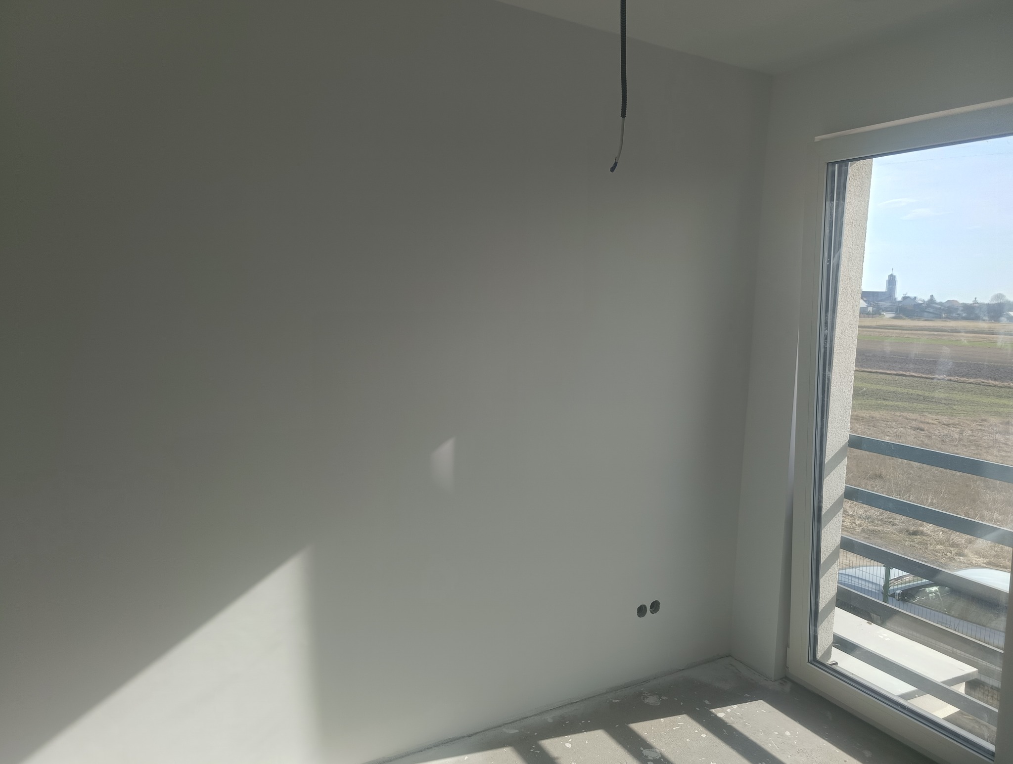Świeżo pomalowana ściana w jasnym odcieniu szarości z widocznymi dwoma otworami na gniazdka elektryczne i zwisającym kablem, balkon z metalową barierką i widokiem na wieś z kościołem w tle.