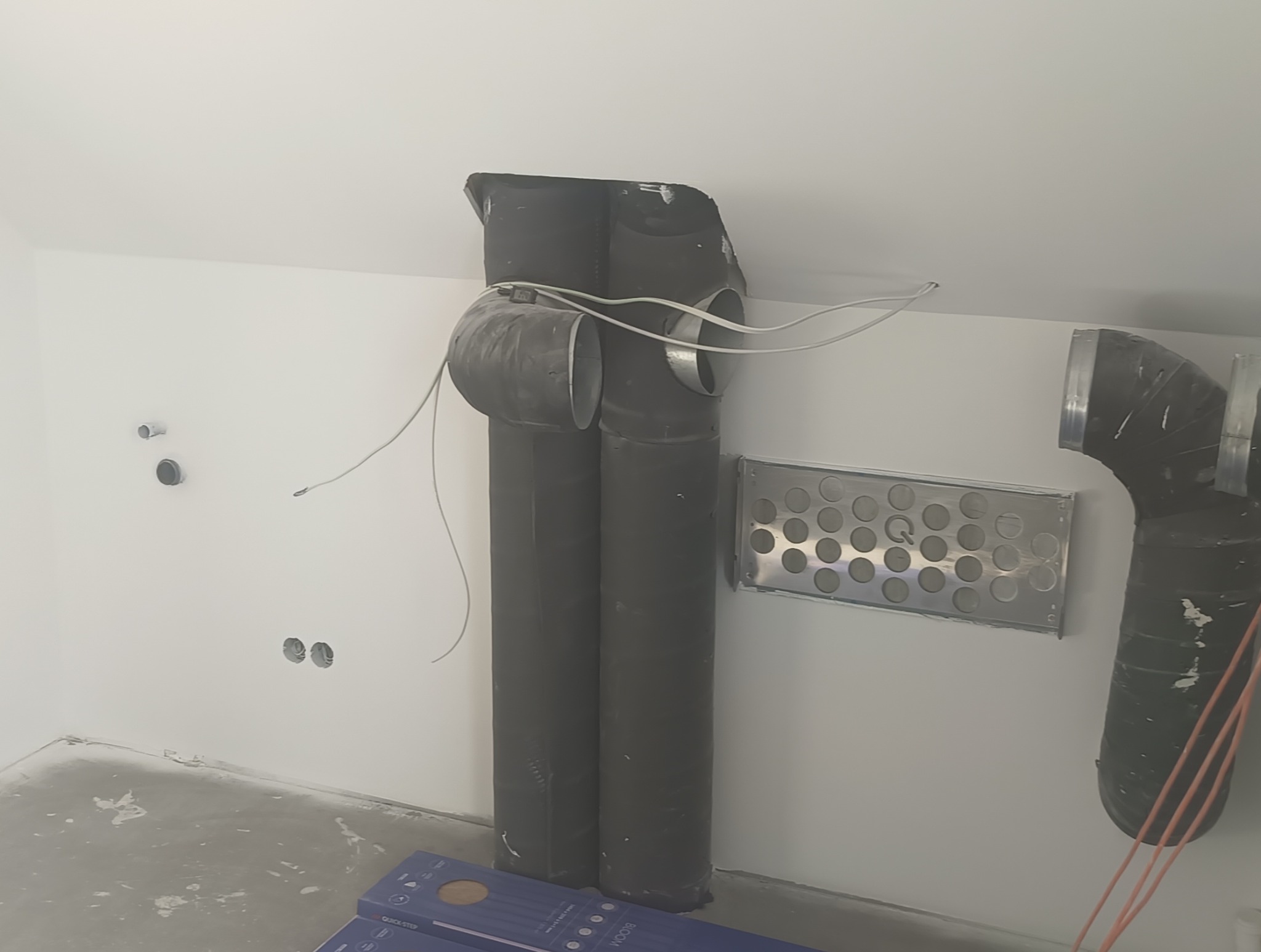 Surowe wykończenie wnętrza z widocznymi rurami wentylacyjnymi, metalową kratką wentylacyjną i wystającymi przewodami elektrycznymi, podłoga częściowo pokryta wylewką, w tle puszki instalacyjne.