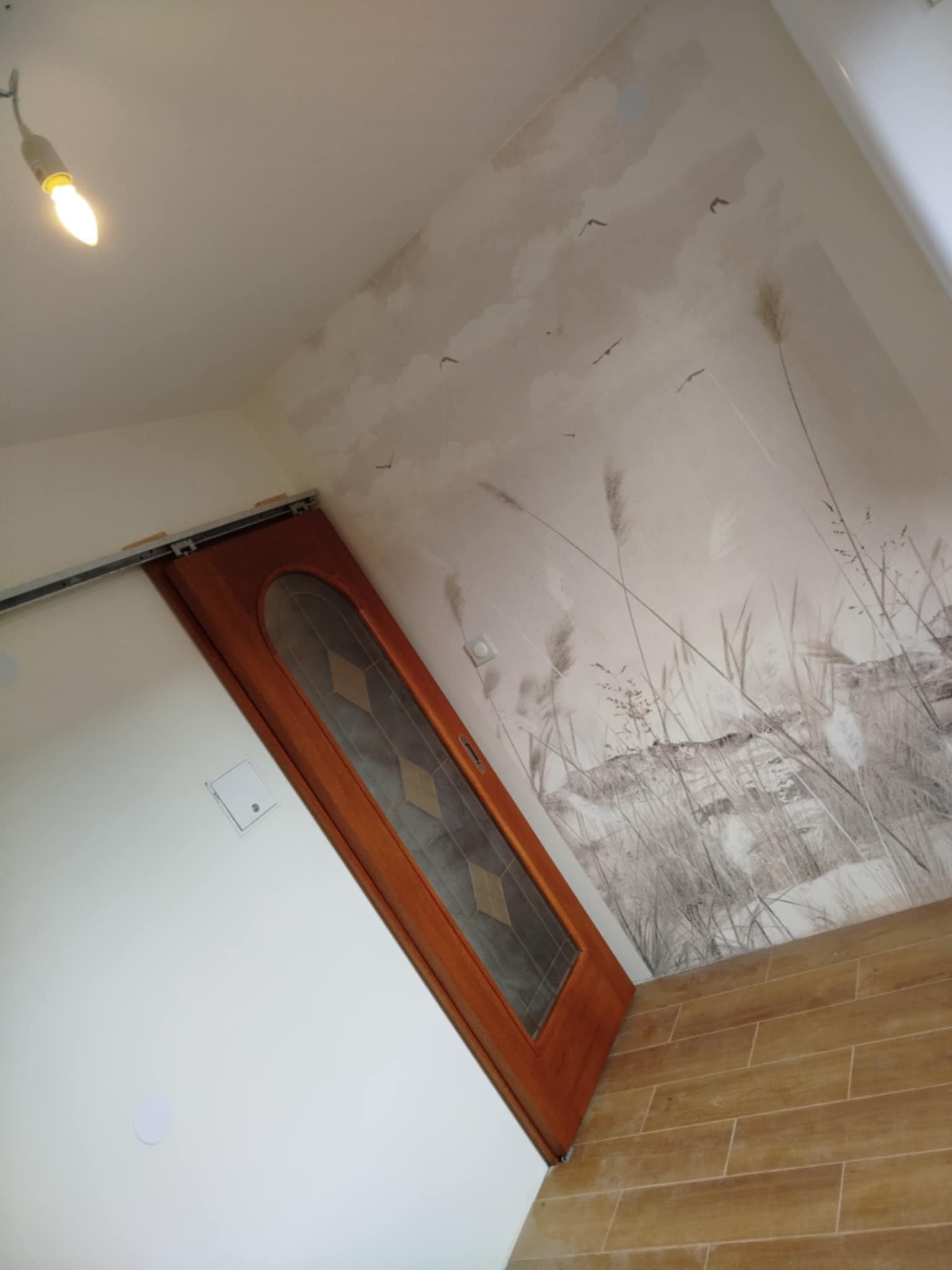 Korytarz po remoncie: drewniana podłoga, drzwi przesuwne z szybą i dekoracyjna tapeta z motywem łąki i ptaków. Widoczna żarówka na suficie.