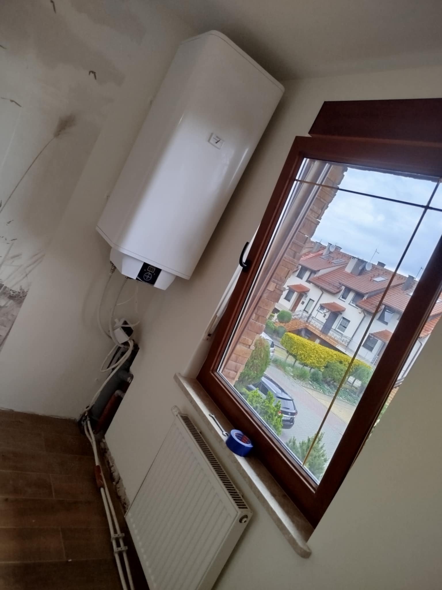 Biały bojler zamontowany na ścianie, widoczne rury i grzejnik. W tle okno z widokiem na osiedle domów. Ściana wymaga wykończenia.