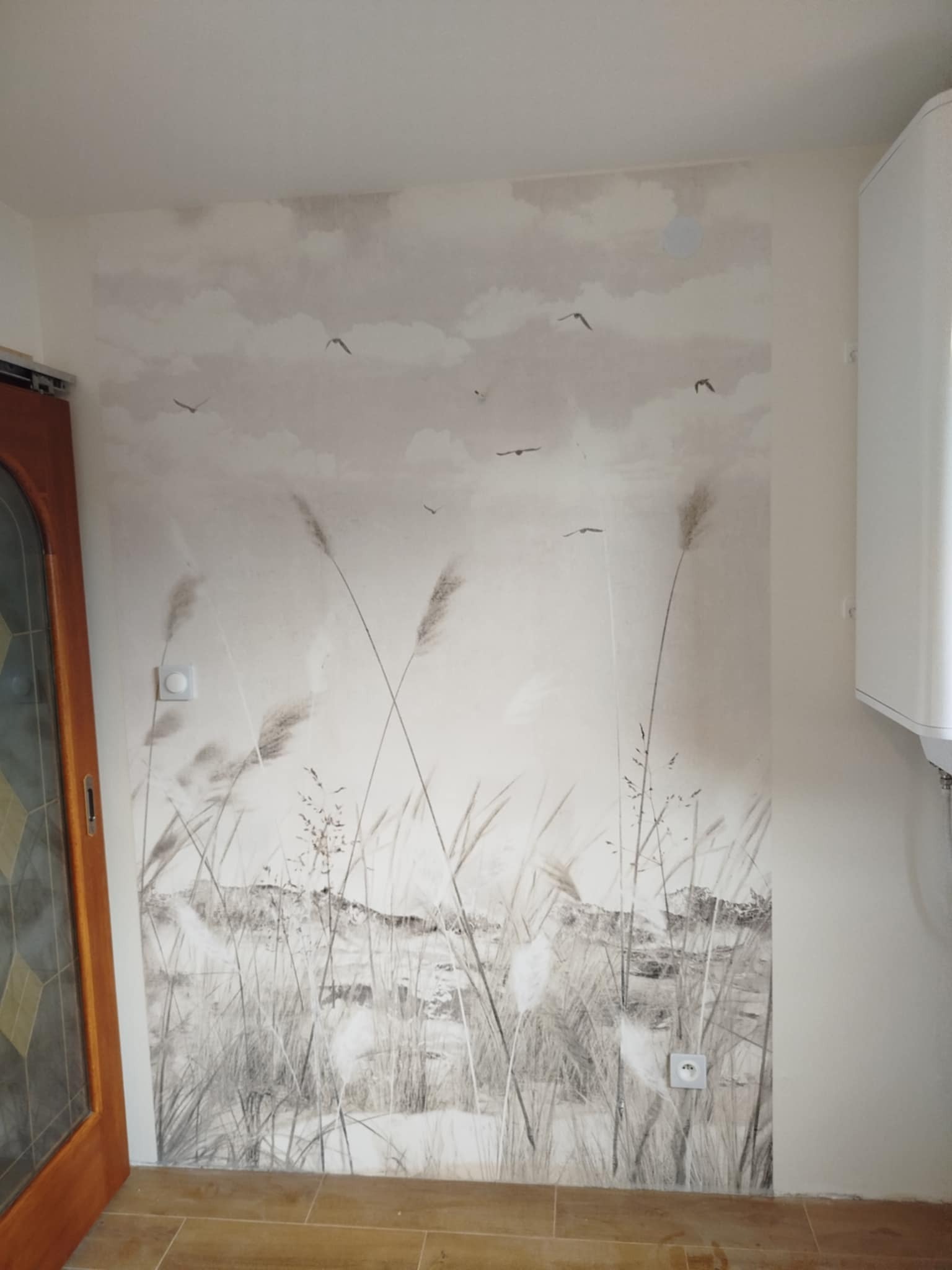 Ściana z fototapetą w odcieniach beżu, przedstawiającą motyw łąki z ptakami. Widoczny fragment drzwi i grzejnik. Na ścianie gniazdko i włącznik.