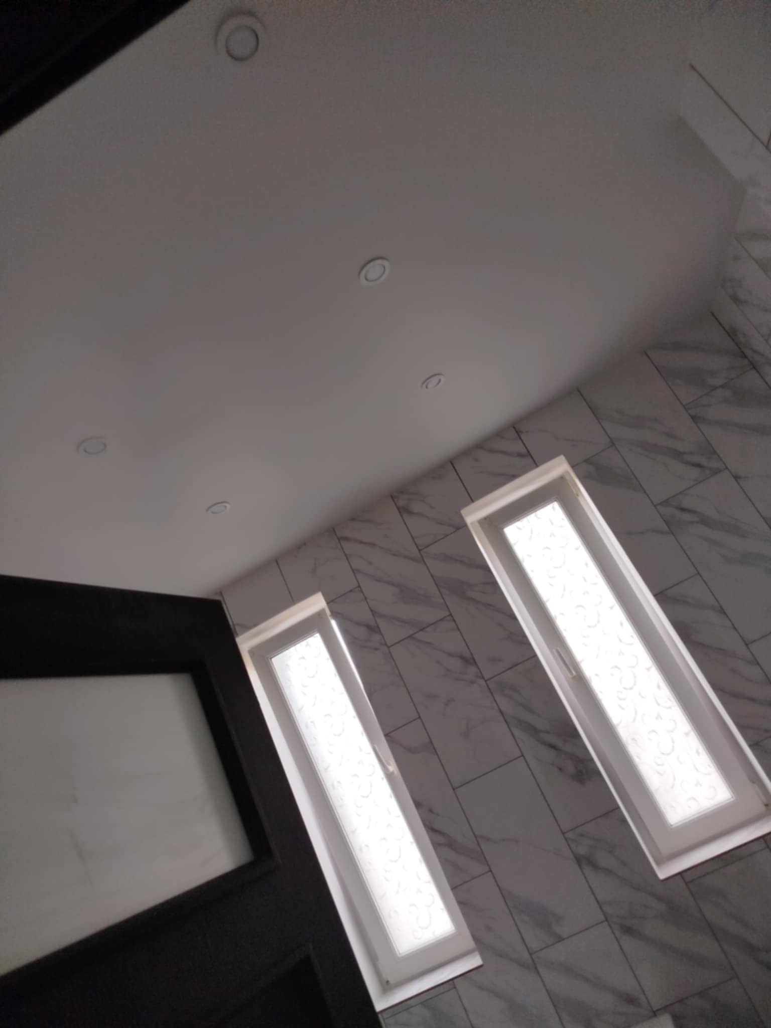 Wykończona łazienka z marmurowymi płytkami, białym sufitem z oświetleniem punktowym i dwoma oknami z ozdobnymi szybami. Ciemne drzwi w lewym dolnym rogu.