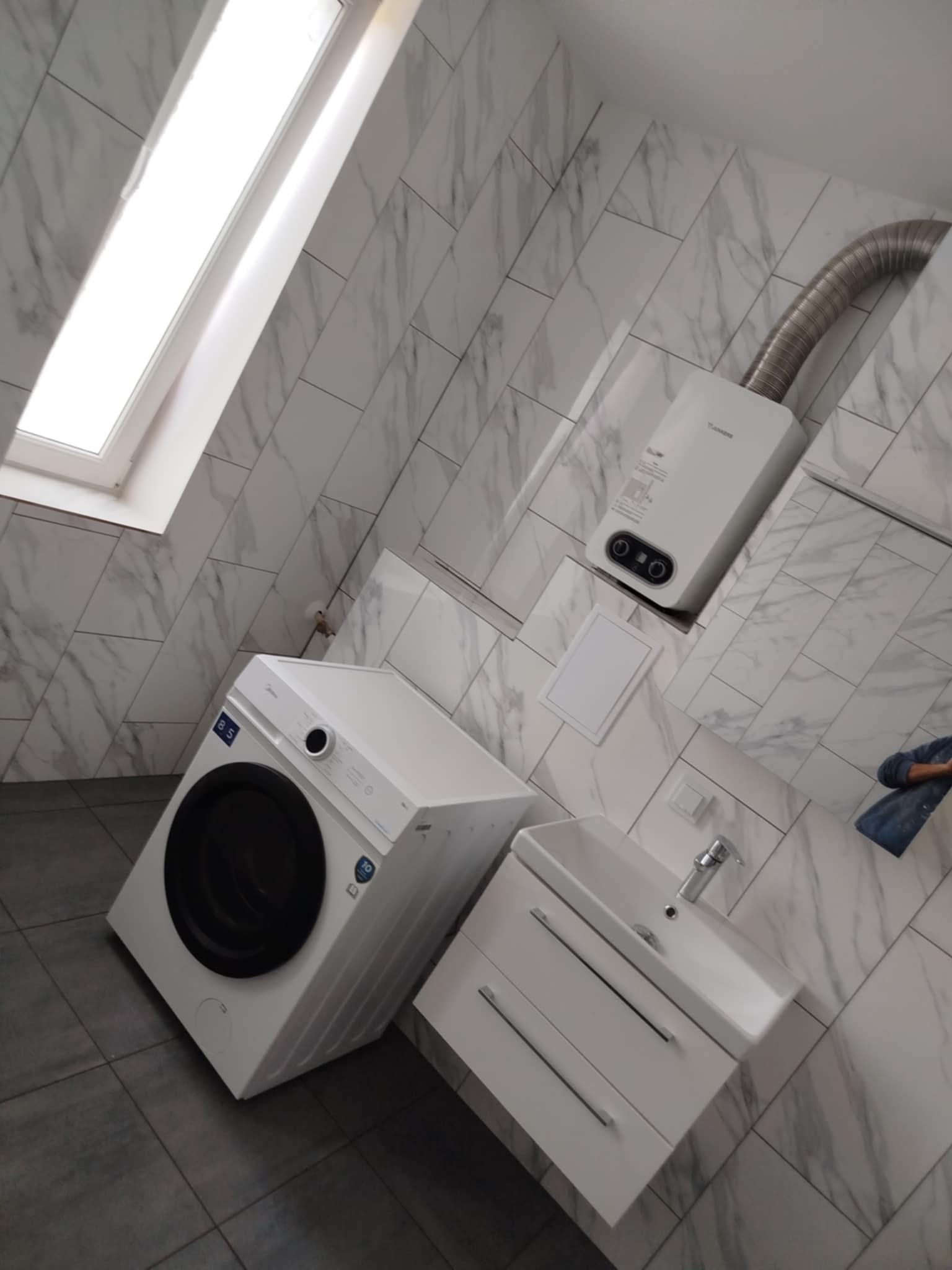 Biała łazienka z marmurowymi płytkami, pralką, umywalką z szafką i bojlerem. Ukośne ujęcie, widoczne okno dachowe i szare płytki podłogowe.